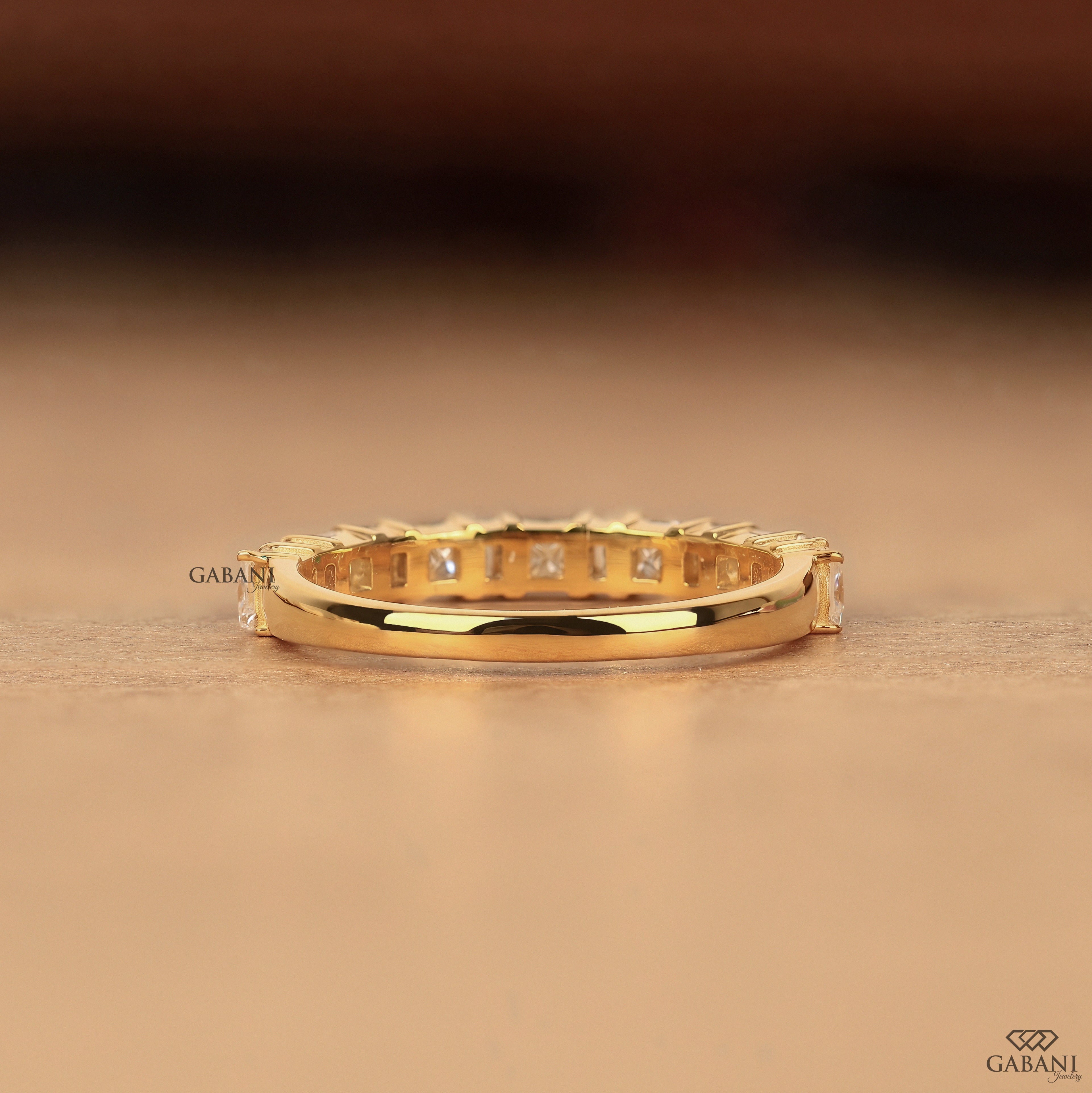 A gold eternity ring with square-cut clear stones set all around, resting on a warm brown surface. The brand name GABANI is visible on the ring and as a logo in the bottom right corner.