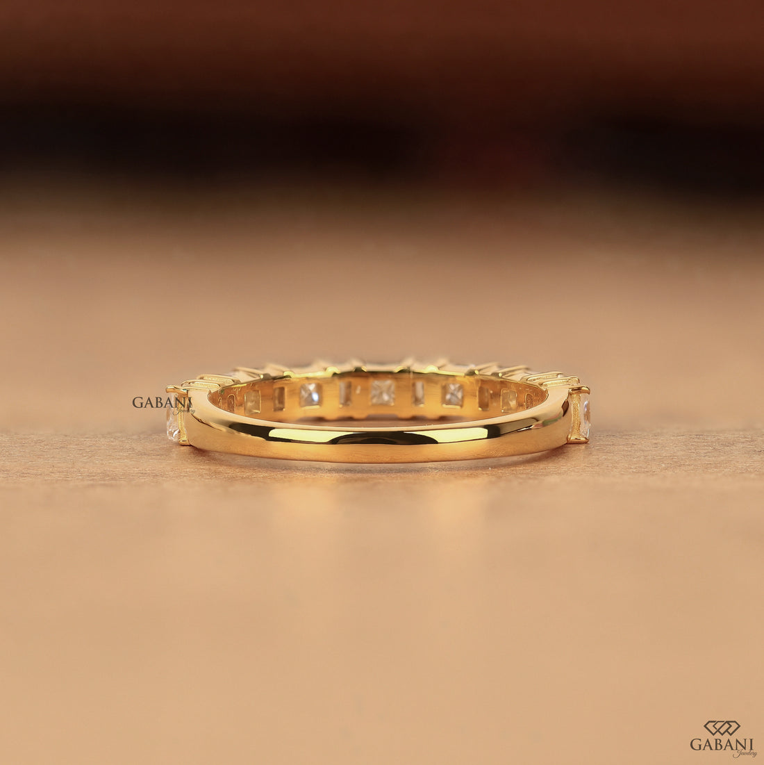 A gold eternity ring with square-cut clear stones set all around, resting on a warm brown surface. The brand name GABANI is visible on the ring and as a logo in the bottom right corner.