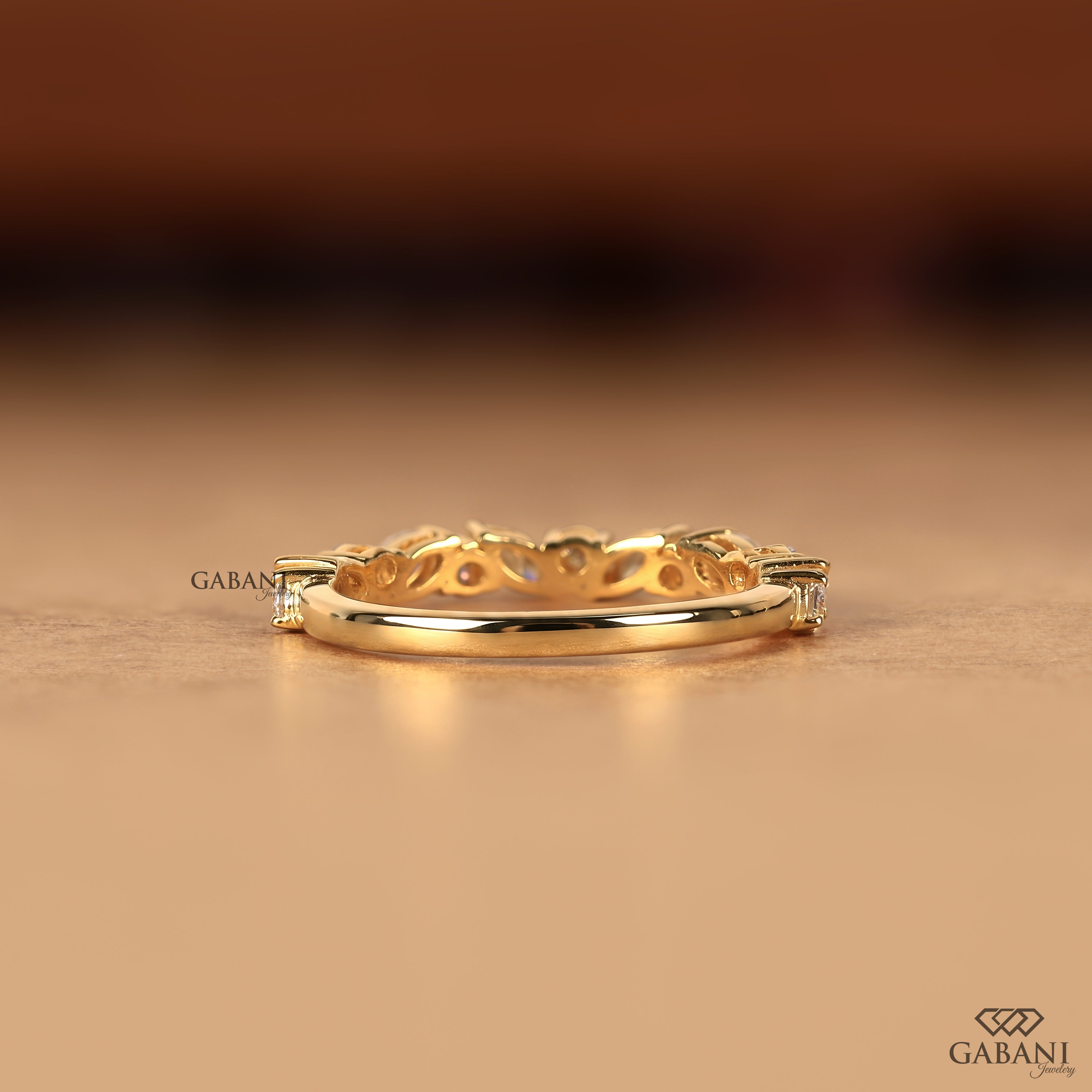 A gold ring with a decorative openwork pattern and small clear gemstones on the sides, photographed on a warm beige surface with a blurred brown background. The brand name GABANI is visible as a watermark and logo.