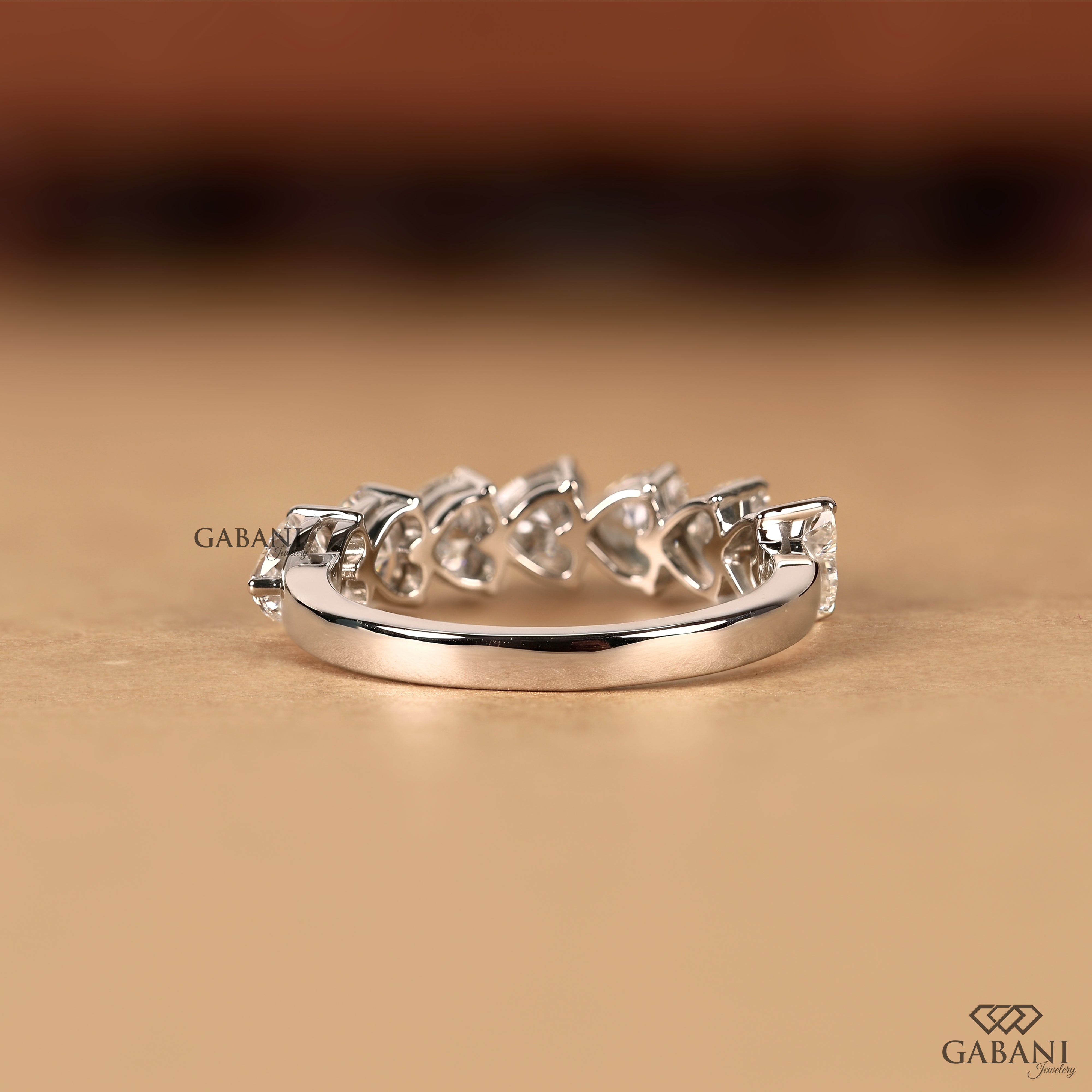 A silver band ring featuring a row of small clear heart-shaped gemstones, photographed on a warm beige surface with a blurred brown background. The brand name GABANI is visible as a watermark and logo.