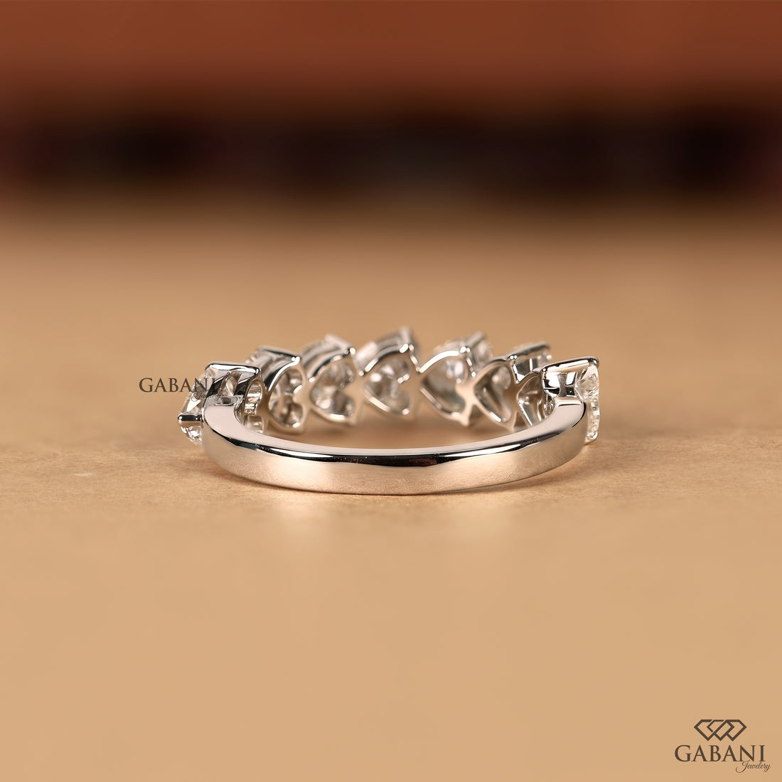 A silver band ring featuring a row of small clear heart-shaped gemstones, photographed on a warm beige surface with a blurred brown background. The brand name GABANI is visible as a watermark and logo.