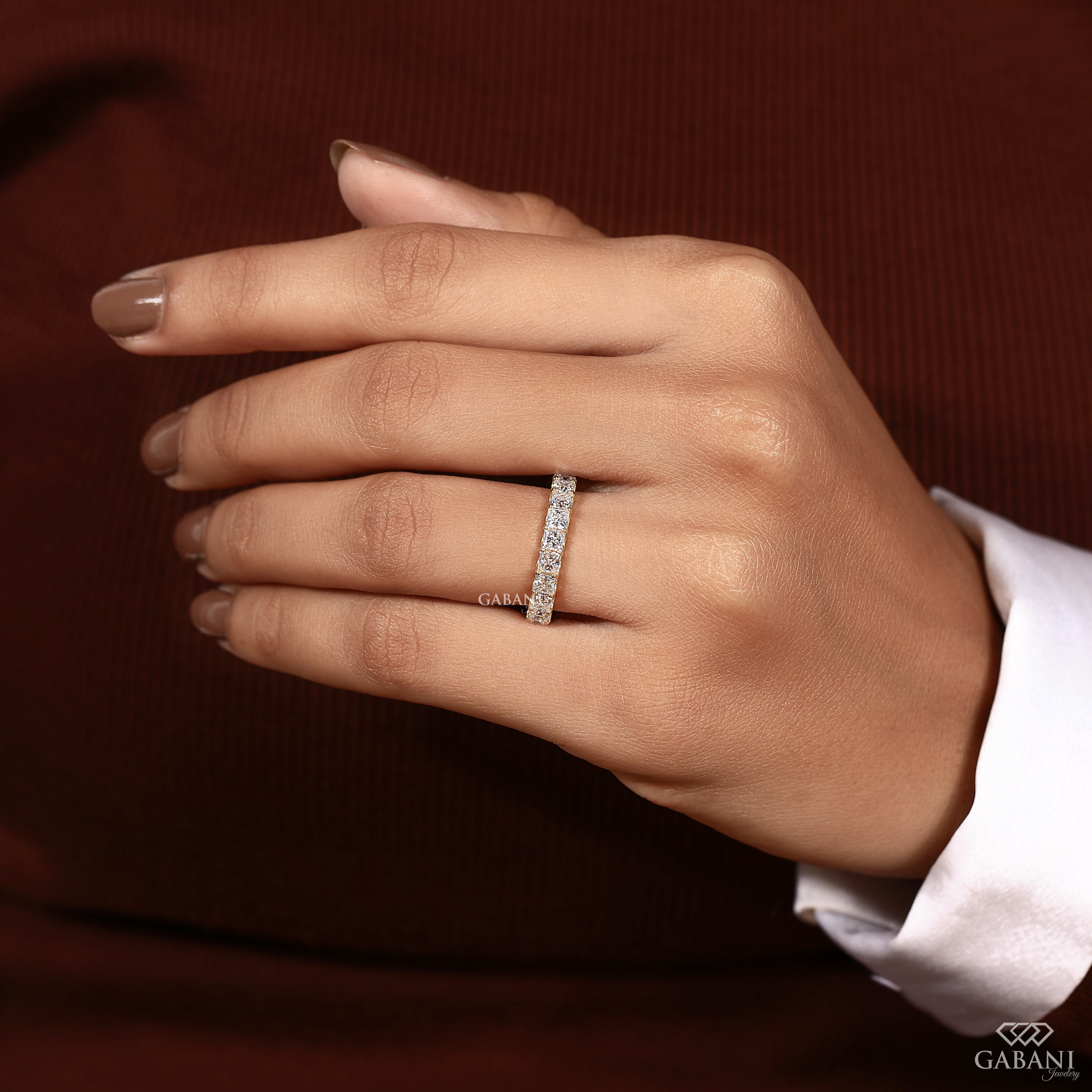 A close-up of a hand wearing a slim diamond band ring on the ring finger, set against a dark brown ribbed background with a white shirt cuff visible.