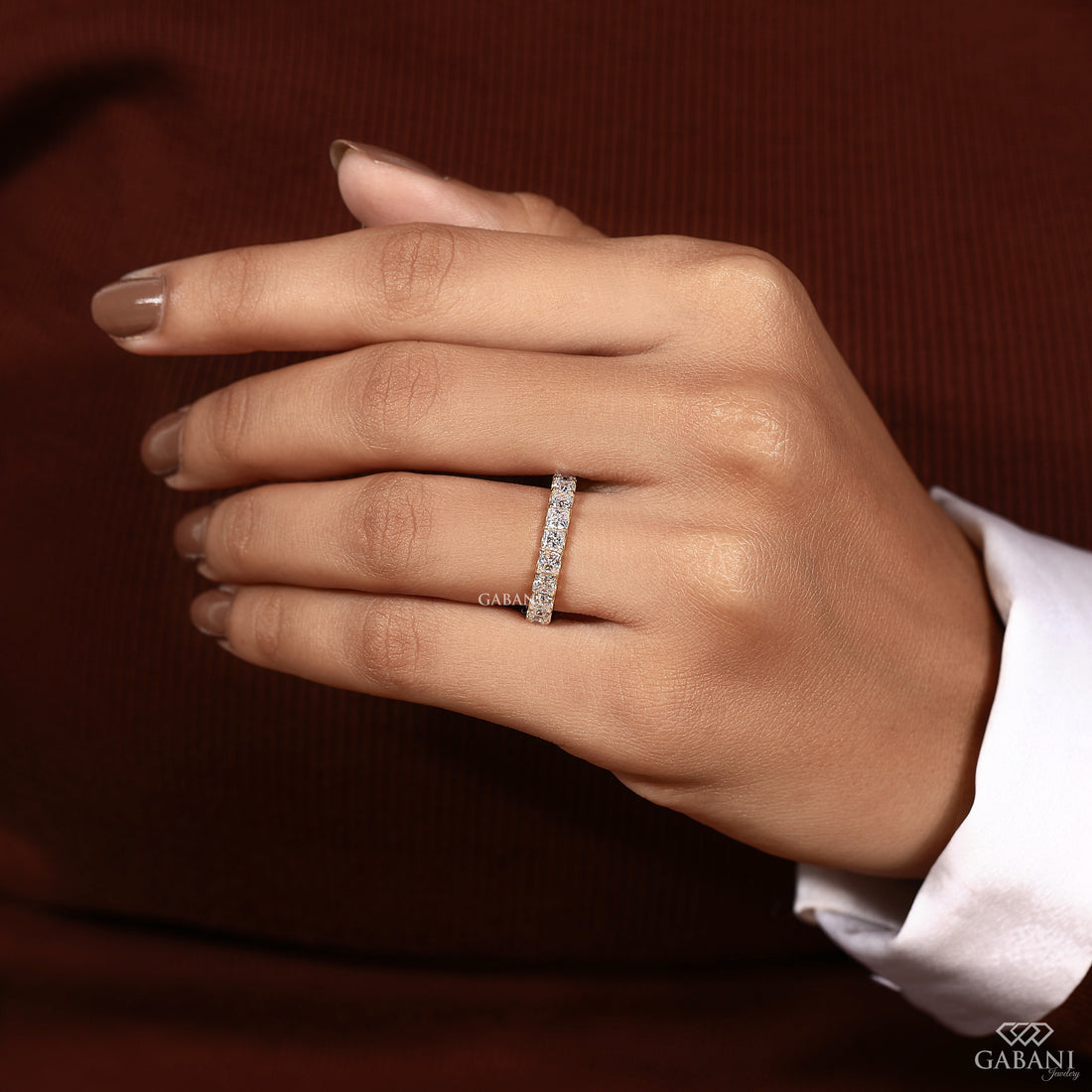 A close-up of a hand wearing a slim diamond band ring on the ring finger, set against a dark brown ribbed background with a white shirt cuff visible.