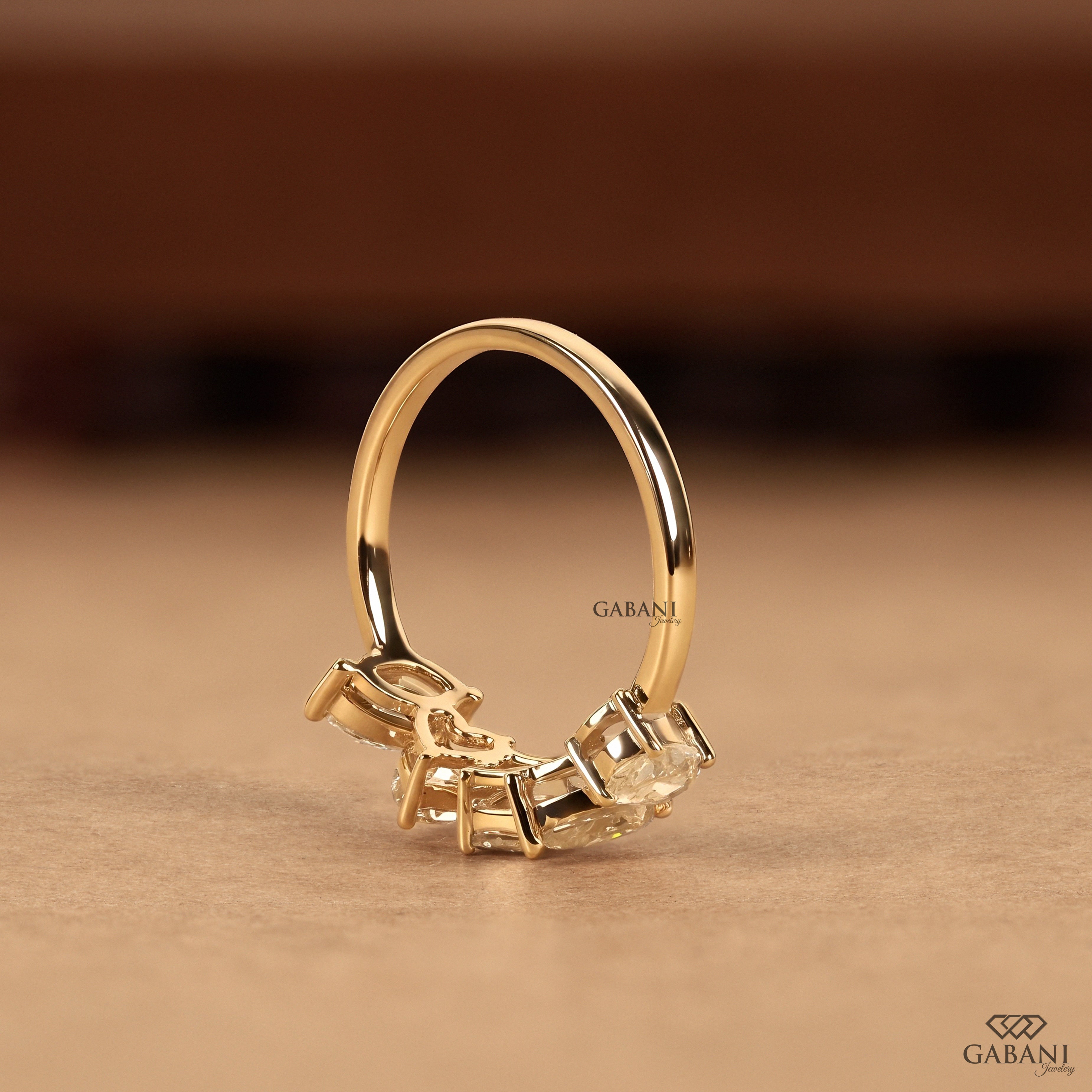 A gold ring with a curved band and a cluster of clear gemstones at the front, photographed on a warm brown surface with a blurred brown background. The brand name GABANI is visible on the ring and in the bottom right corner.