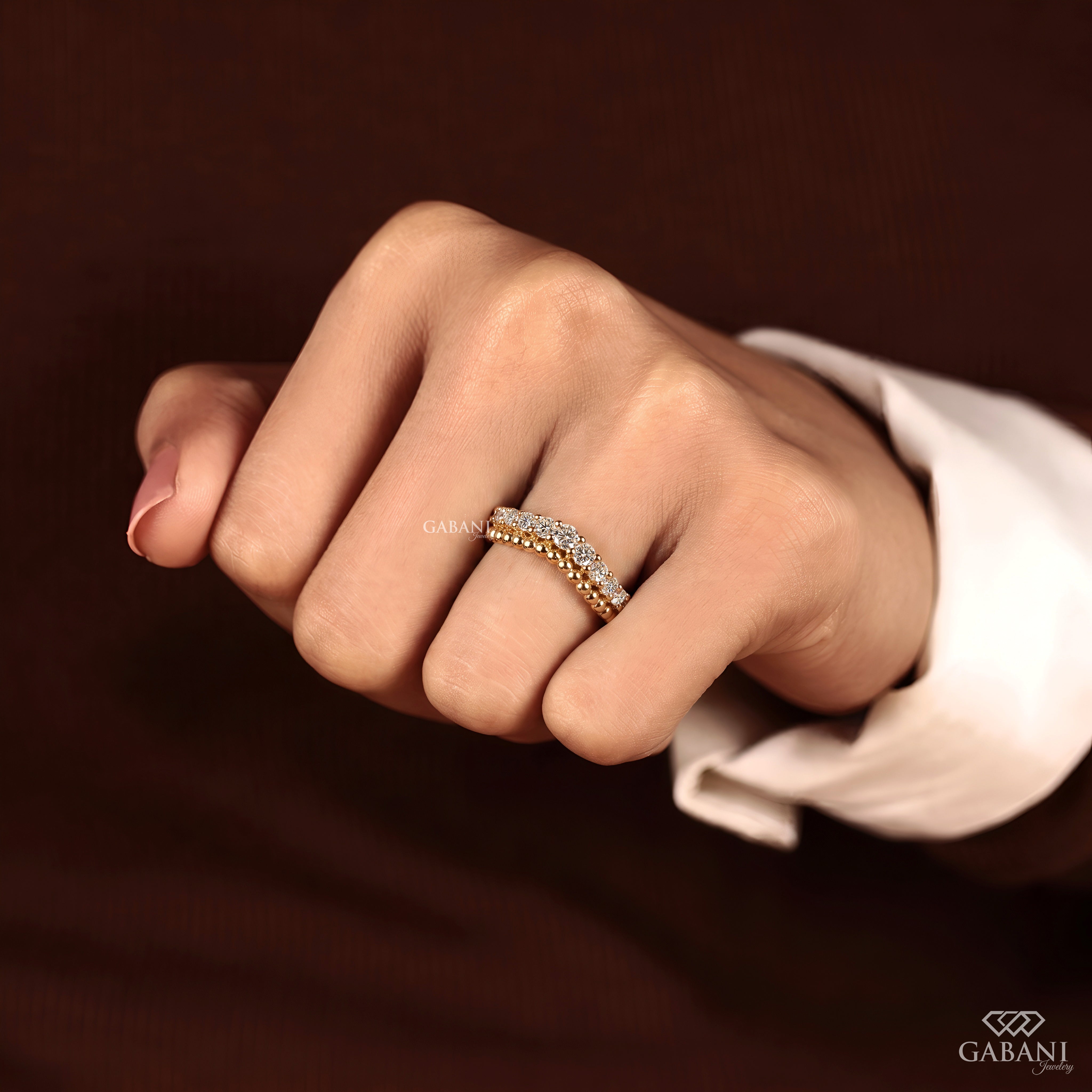 A close-up of a hand wearing a delicate gold band ring with a row of small clear stones, set against a dark brown background with a white shirt cuff visible.