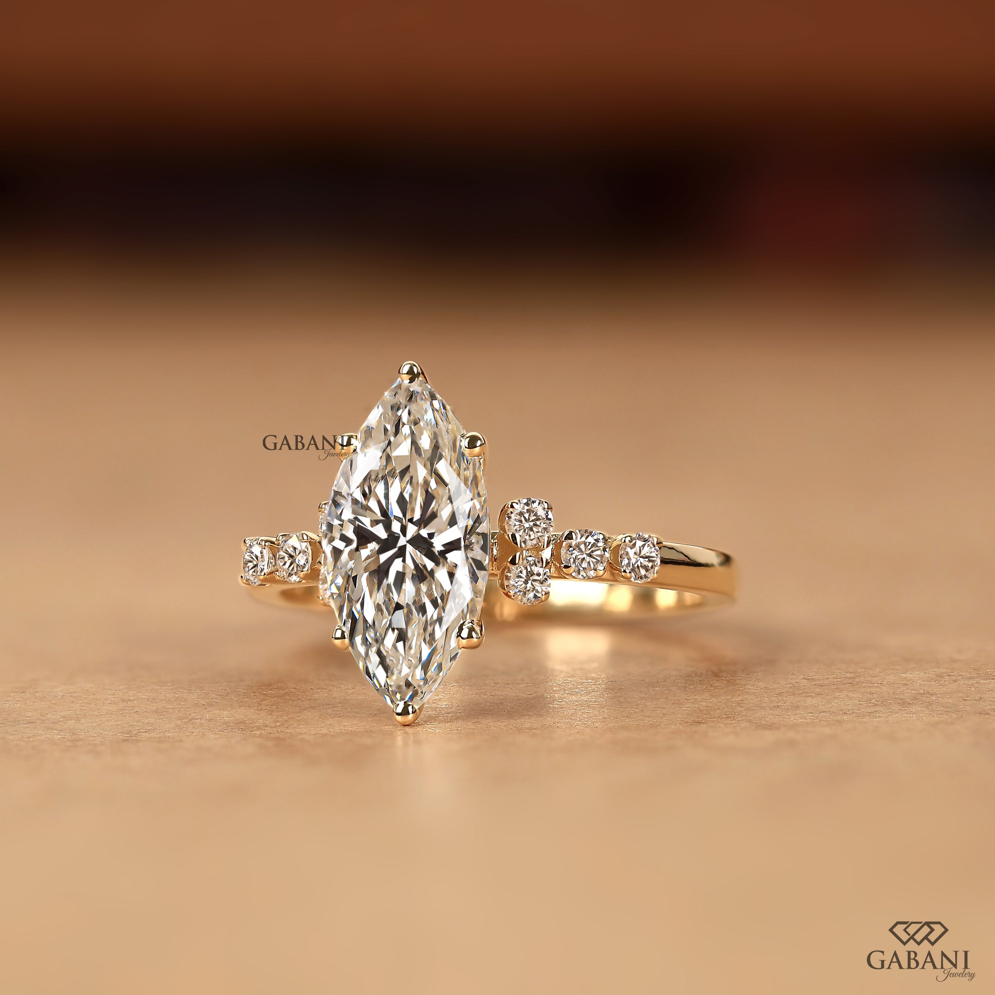 A gold ring featuring a large marquise-cut clear gemstone in a prong setting, with smaller round-cut stones on each side of the band, photographed on a warm beige surface with a blurred brown background. The brand name GABANI is visible as a watermark.