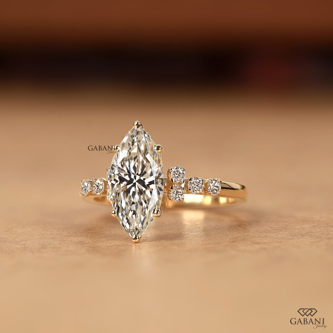 A gold ring featuring a large marquise-cut clear gemstone in a prong setting, with smaller round-cut stones on each side of the band, photographed on a warm beige surface with a blurred brown background. The brand name GABANI is visible as a watermark.