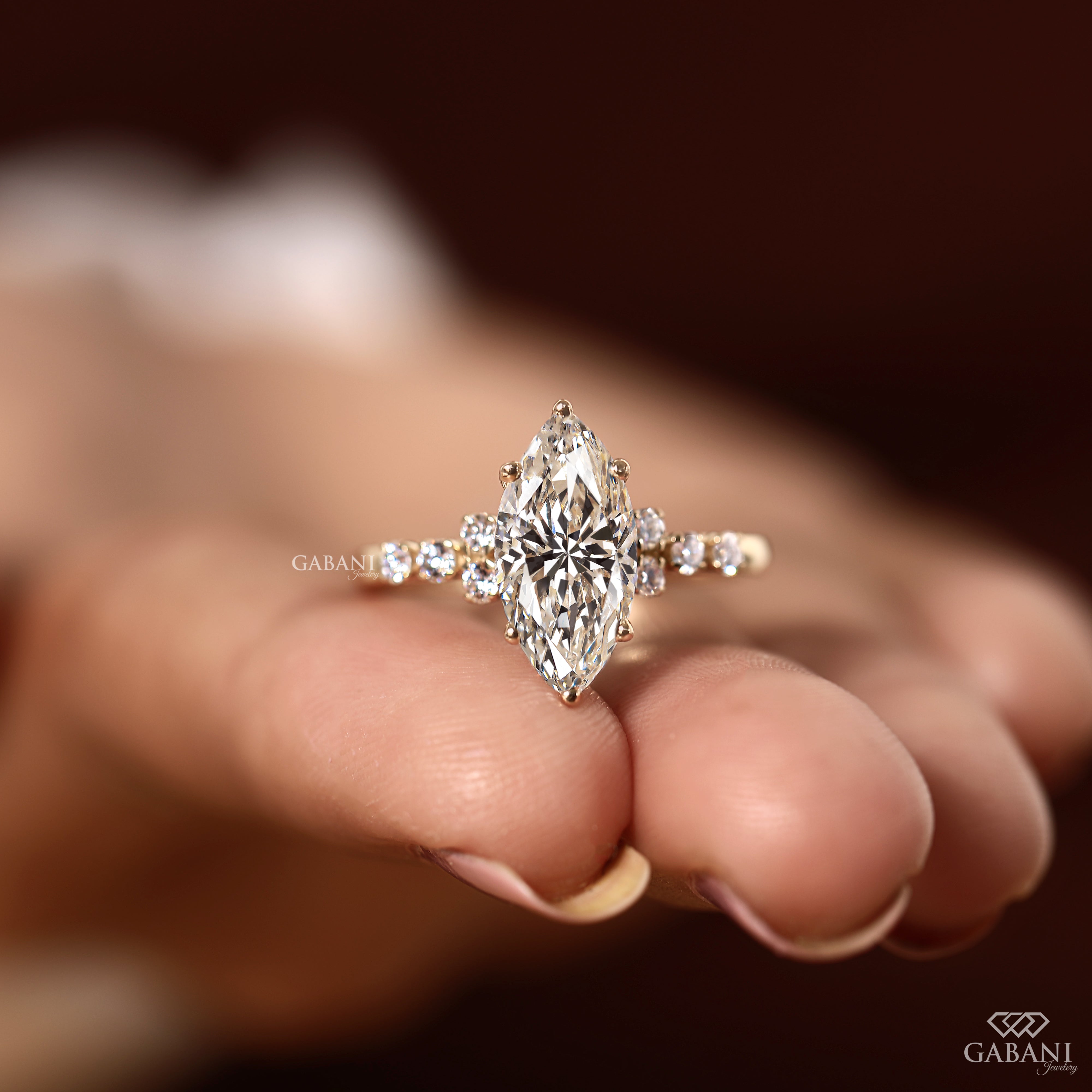 A close-up of a hand holding a gold ring with a marquise-cut diamond center stone and smaller diamonds on the band, set against a dark brown blurred background.