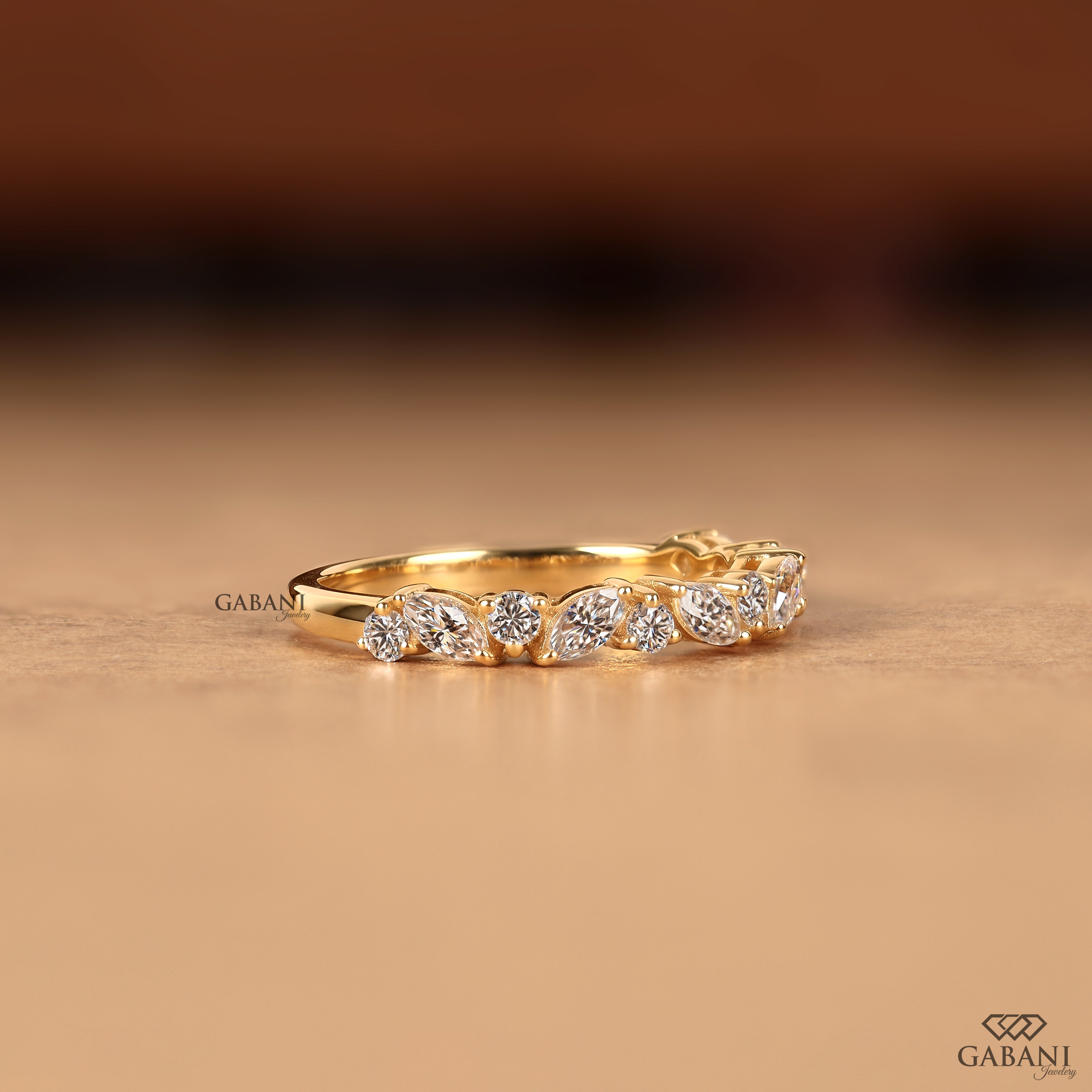 A yellow gold ring featuring a row of marquise-cut and round brilliant-cut diamonds, displayed on a warm beige surface with a blurred brown background. The brand name 'GABANI' is visible as a watermark on the left side of the ring.