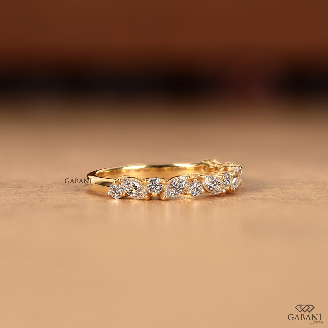 A yellow gold ring featuring a row of marquise-cut and round brilliant-cut diamonds, displayed on a warm beige surface with a blurred brown background. The brand name 'GABANI' is visible as a watermark on the left side of the ring.