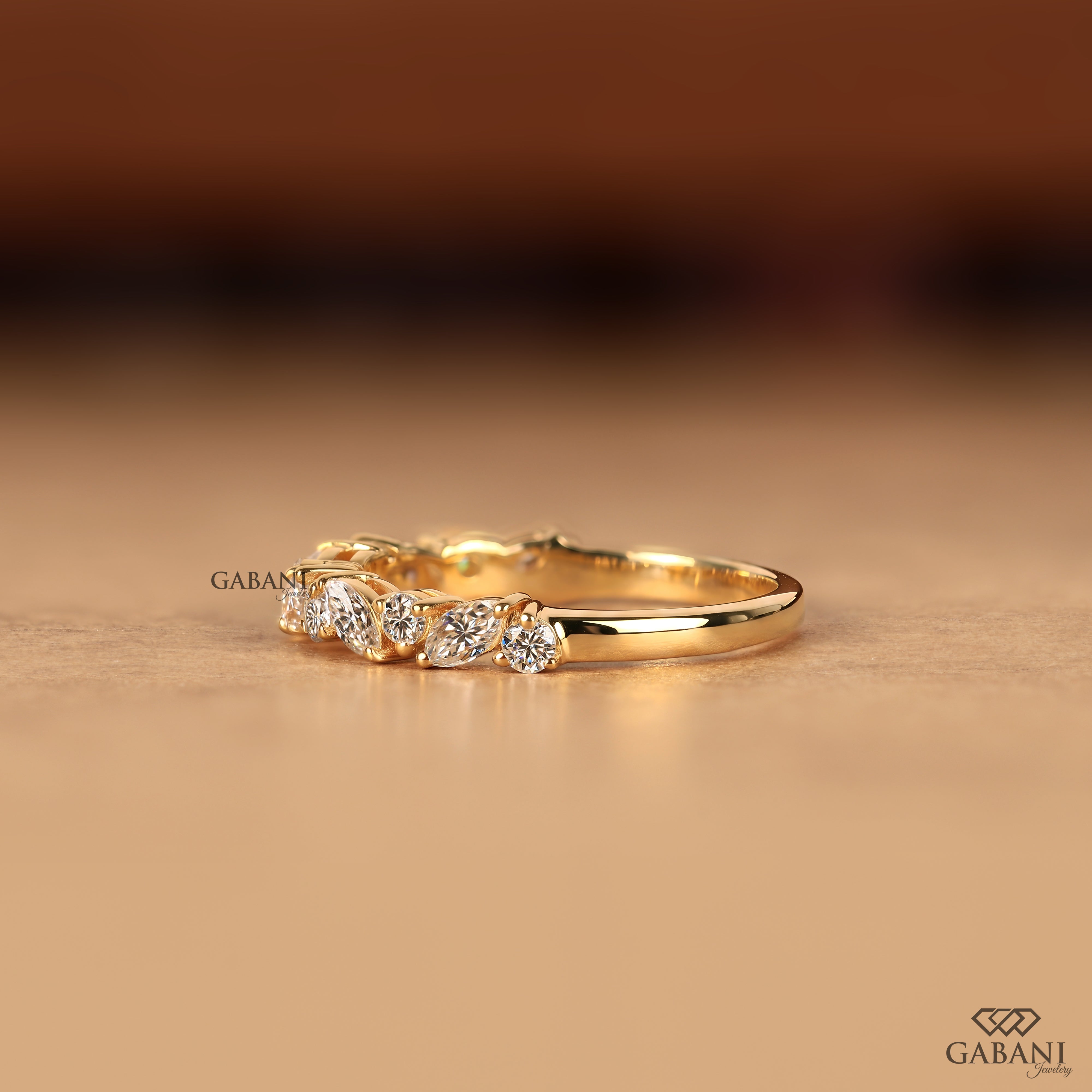 A gold band ring featuring a row of small clear gemstones, photographed on a warm beige surface with a blurred brown background. The brand name GABANI is visible as a watermark and in the bottom right corner.