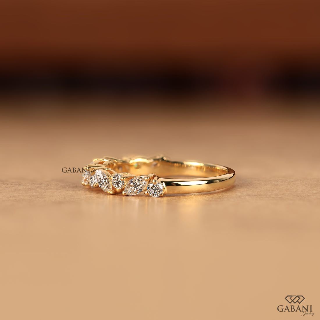 A gold band ring featuring a row of small clear gemstones, photographed on a warm beige surface with a blurred brown background. The brand name GABANI is visible as a watermark and in the bottom right corner.