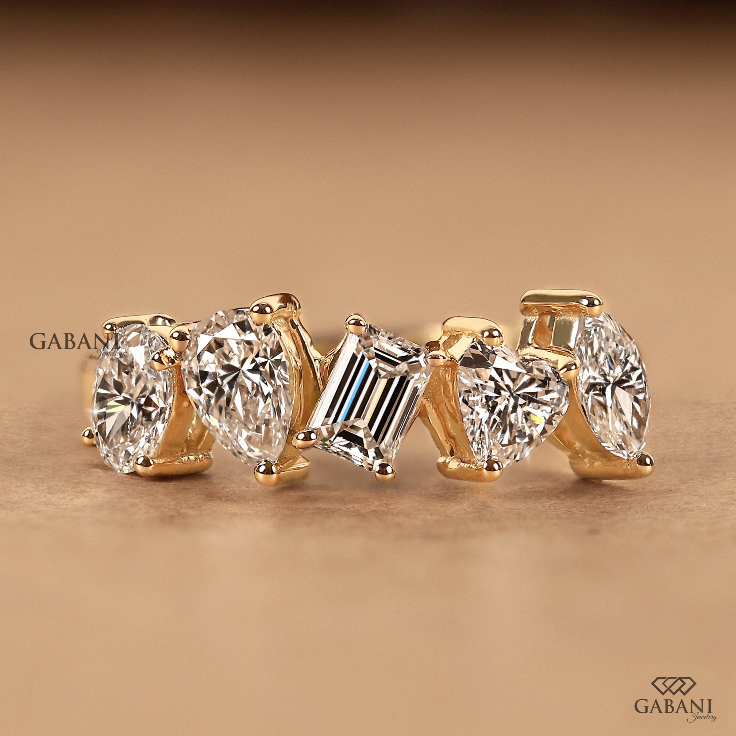 A gold ring featuring five clear gemstones in different cuts: four oval diamonds and one baguette diamond, displayed on a warm brown background with Gabani branding.