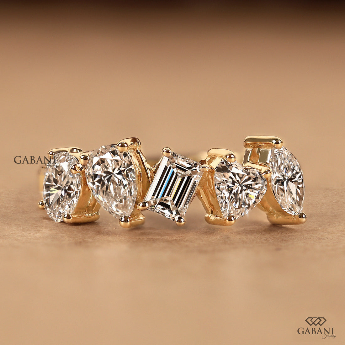 A gold ring featuring five clear gemstones in different cuts: four oval diamonds and one baguette diamond, displayed on a warm brown background with Gabani branding.