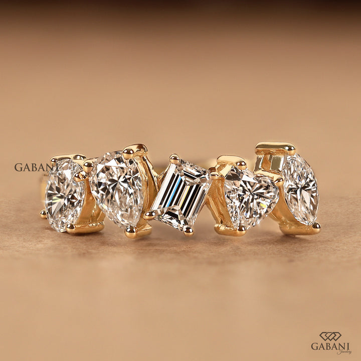 A gold ring featuring five clear gemstones in different cuts: four oval diamonds and one baguette diamond, displayed on a warm brown background with Gabani branding.