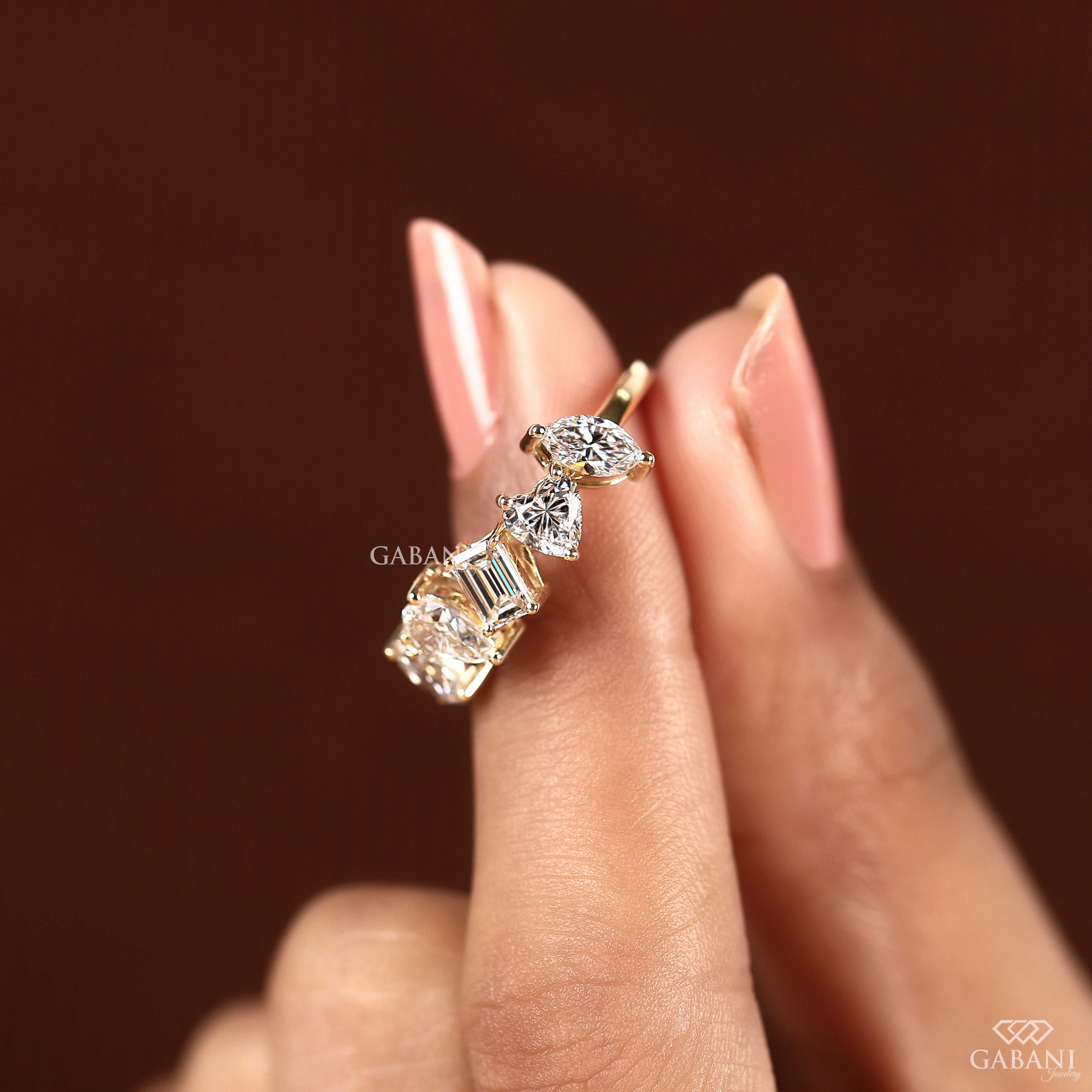A gold ring with a row of clear baguette and marquise-cut gemstones, shown worn on a person's finger against a dark brown background. The brand name 'GABANI' is visible as a watermark.
