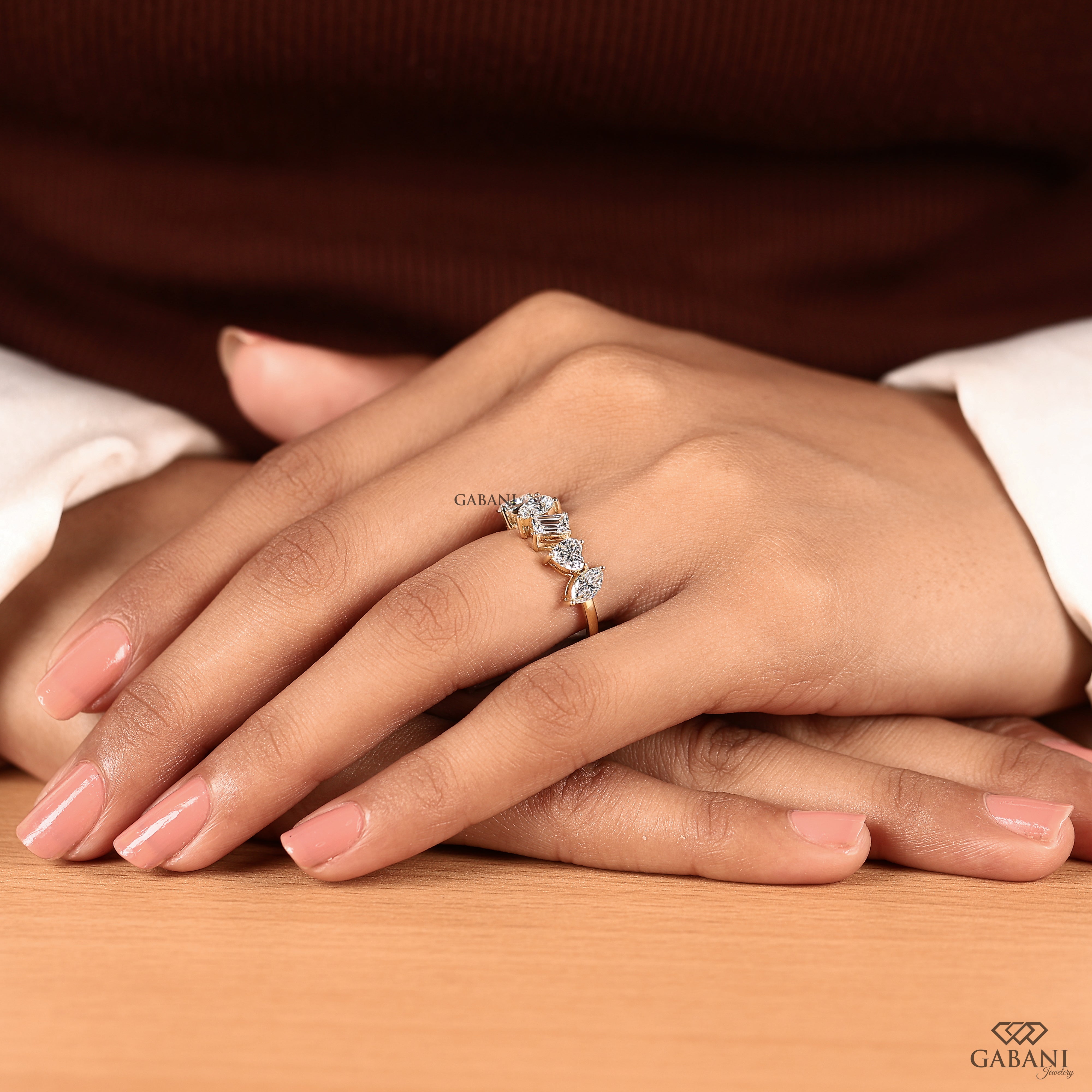 A close-up of a person's hands resting on a wooden surface, wearing a delicate gold ring adorned with small clear marquise-cut gemstones. The background features a dark brown ribbed sweater and a white shirt cuff.