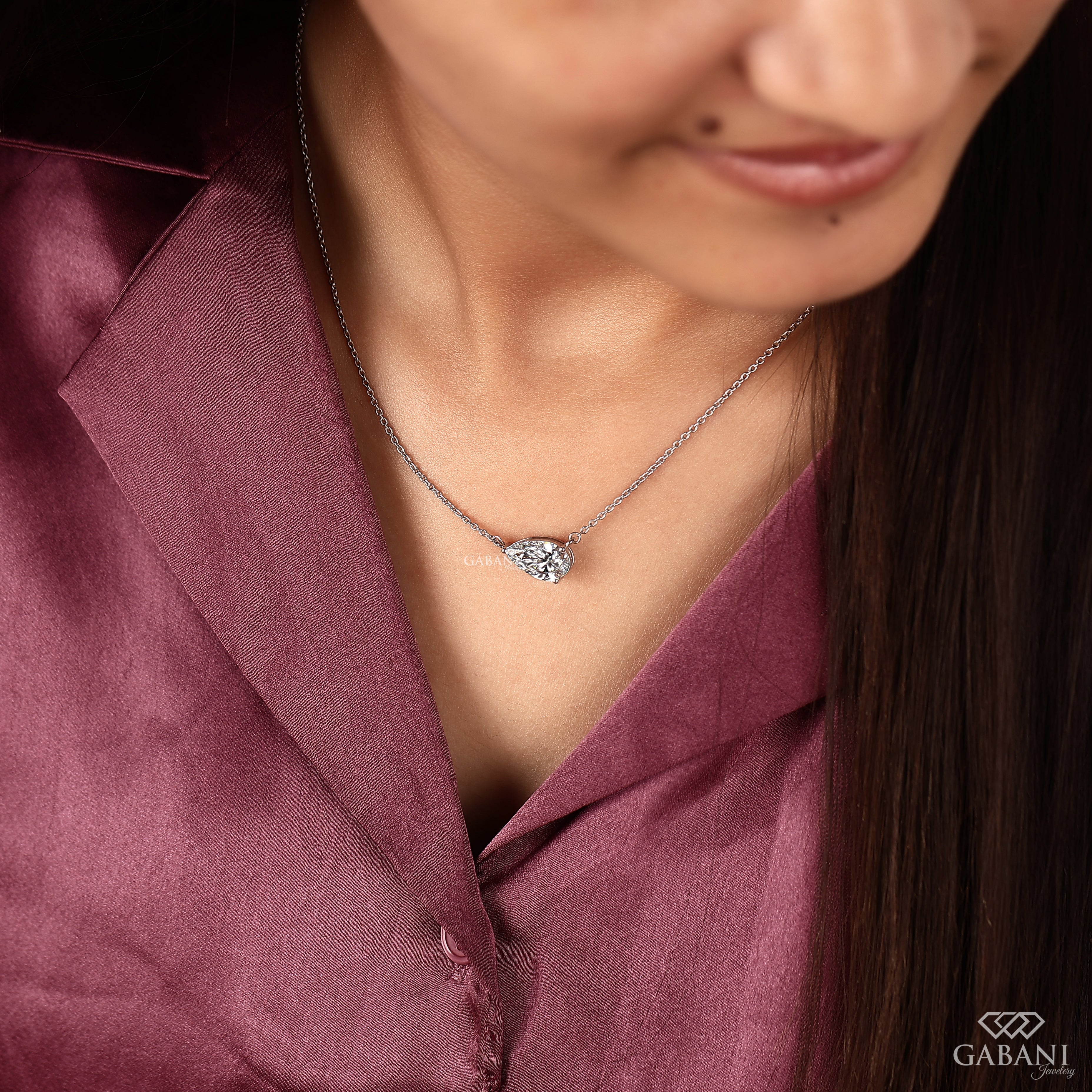 A close-up lifestyle shot of a woman wearing a delicate silver chain necklace with a small, sparkling heart-shaped crystal pendant. She is wearing a satin burgundy blouse, and the brand logo 'GABANI JEWELLERY' is visible in the bottom right corner.