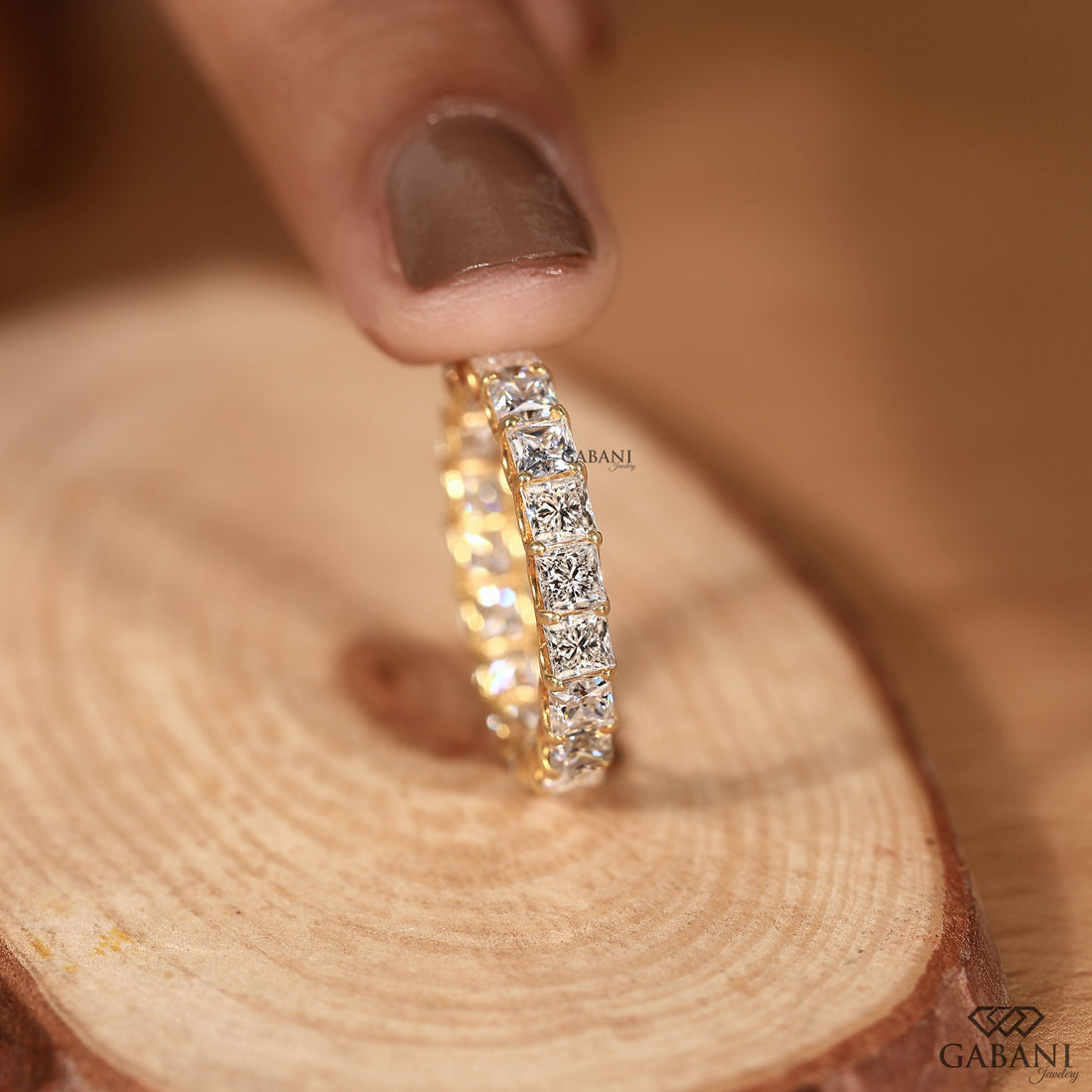 A gold eternity ring set with clear round-cut gemstones, held by a finger over a rustic wood slice. The brand name 'GABANI' is visible as a watermark and logo.