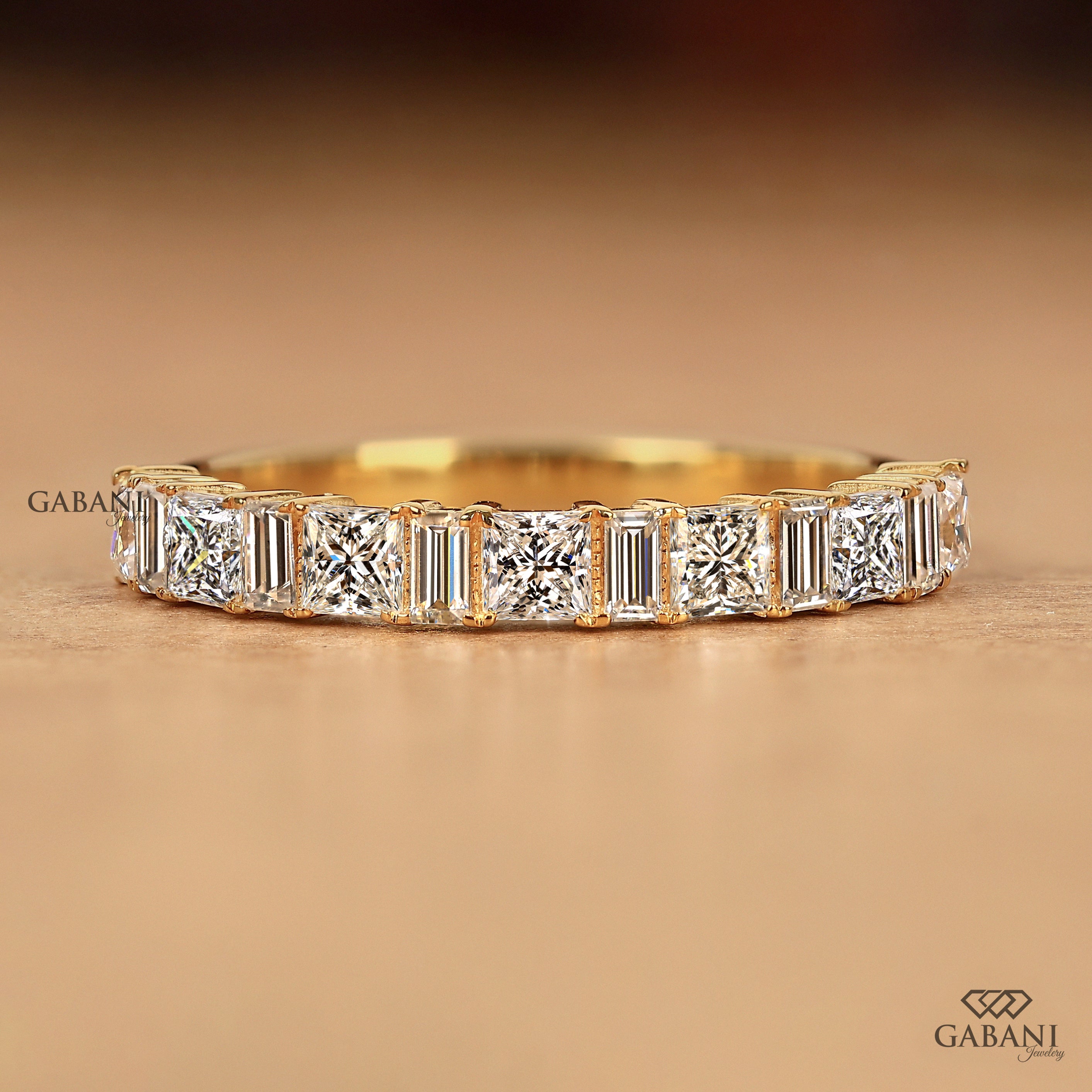 A yellow gold eternity ring featuring a row of baguette and round-cut diamonds, displayed on a warm brown textured surface with the Gabani logo.