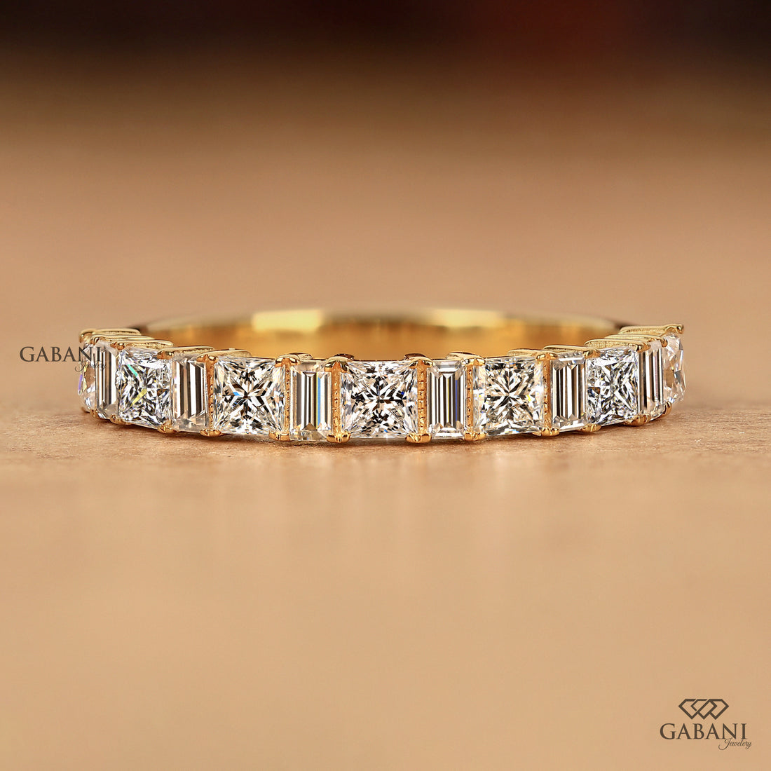 A yellow gold eternity ring featuring a row of baguette and round-cut diamonds, displayed on a warm brown textured surface with the Gabani logo.