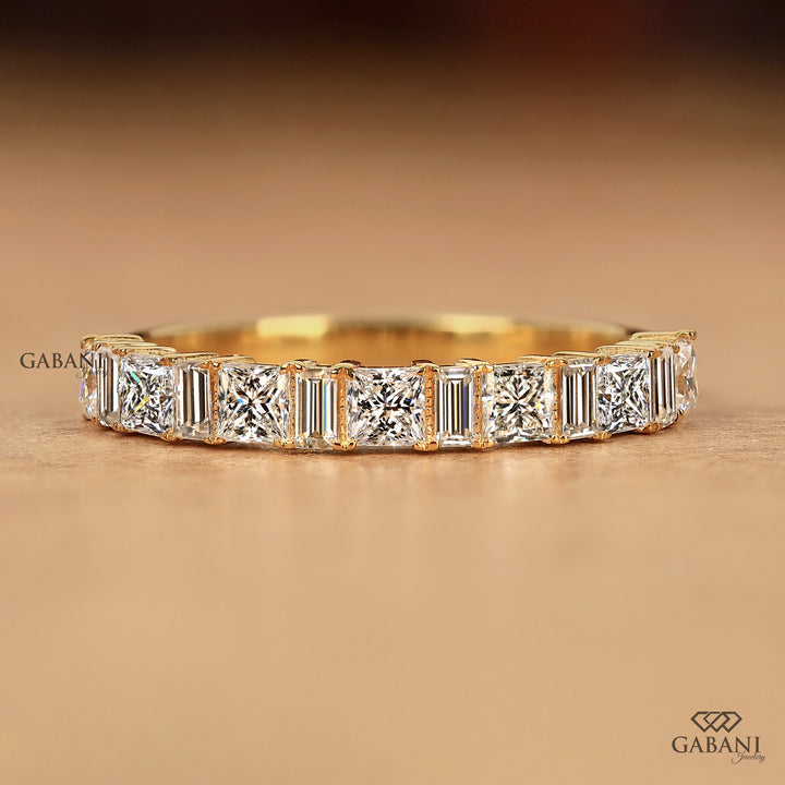 A yellow gold eternity ring featuring a row of baguette and round-cut diamonds, displayed on a warm brown textured surface with the Gabani logo.
