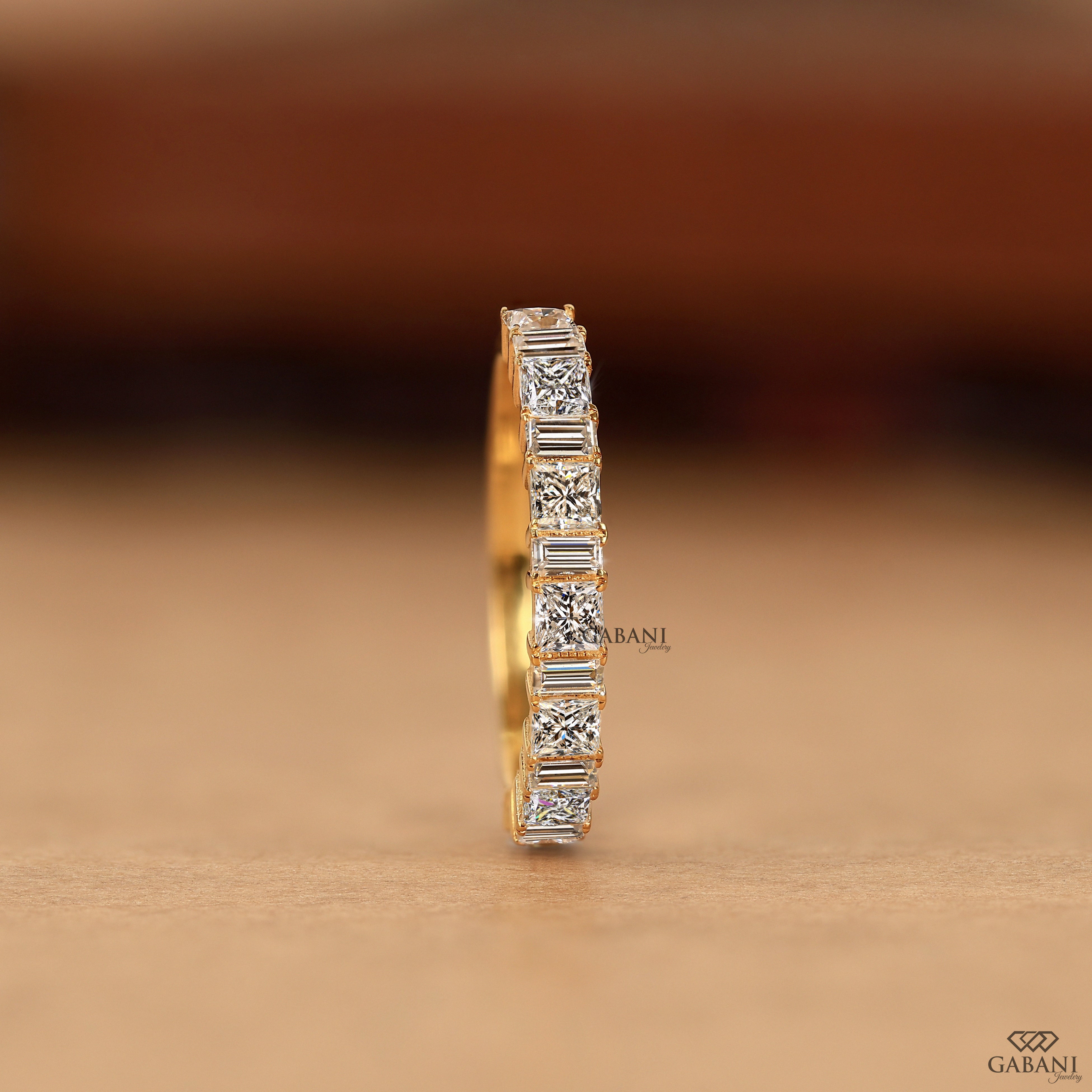 A single yellow gold eternity ring featuring a row of baguette-cut clear gemstones, shown standing upright on a neutral brown surface with a blurred brown background.