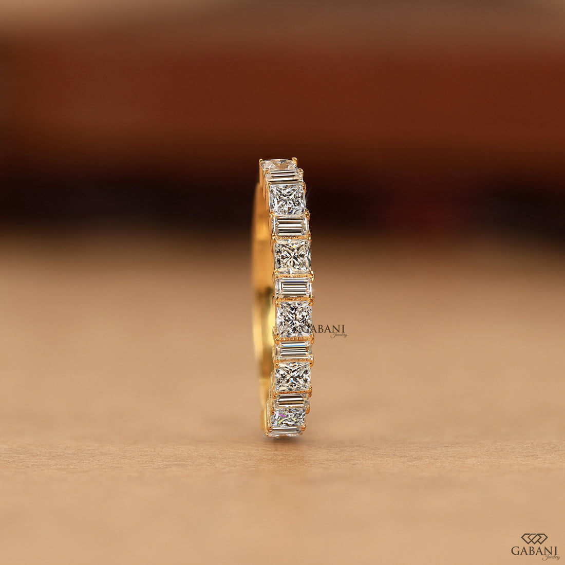 A single yellow gold eternity ring featuring a row of baguette-cut clear gemstones, shown standing upright on a neutral brown surface with a blurred brown background.