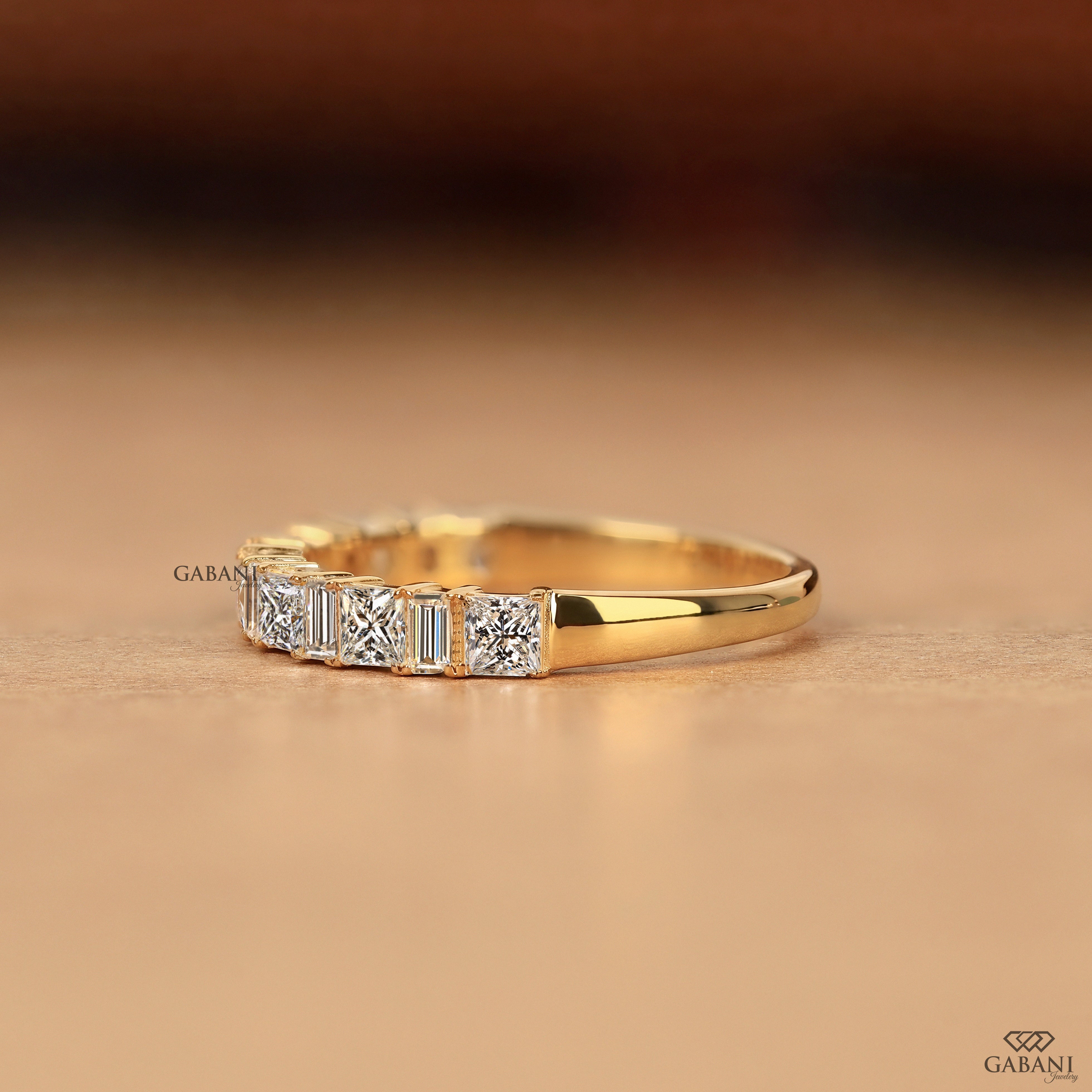 A yellow gold ring featuring a row of baguette-cut and round-cut clear gemstones, displayed on a warm beige surface with a blurred dark brown background.