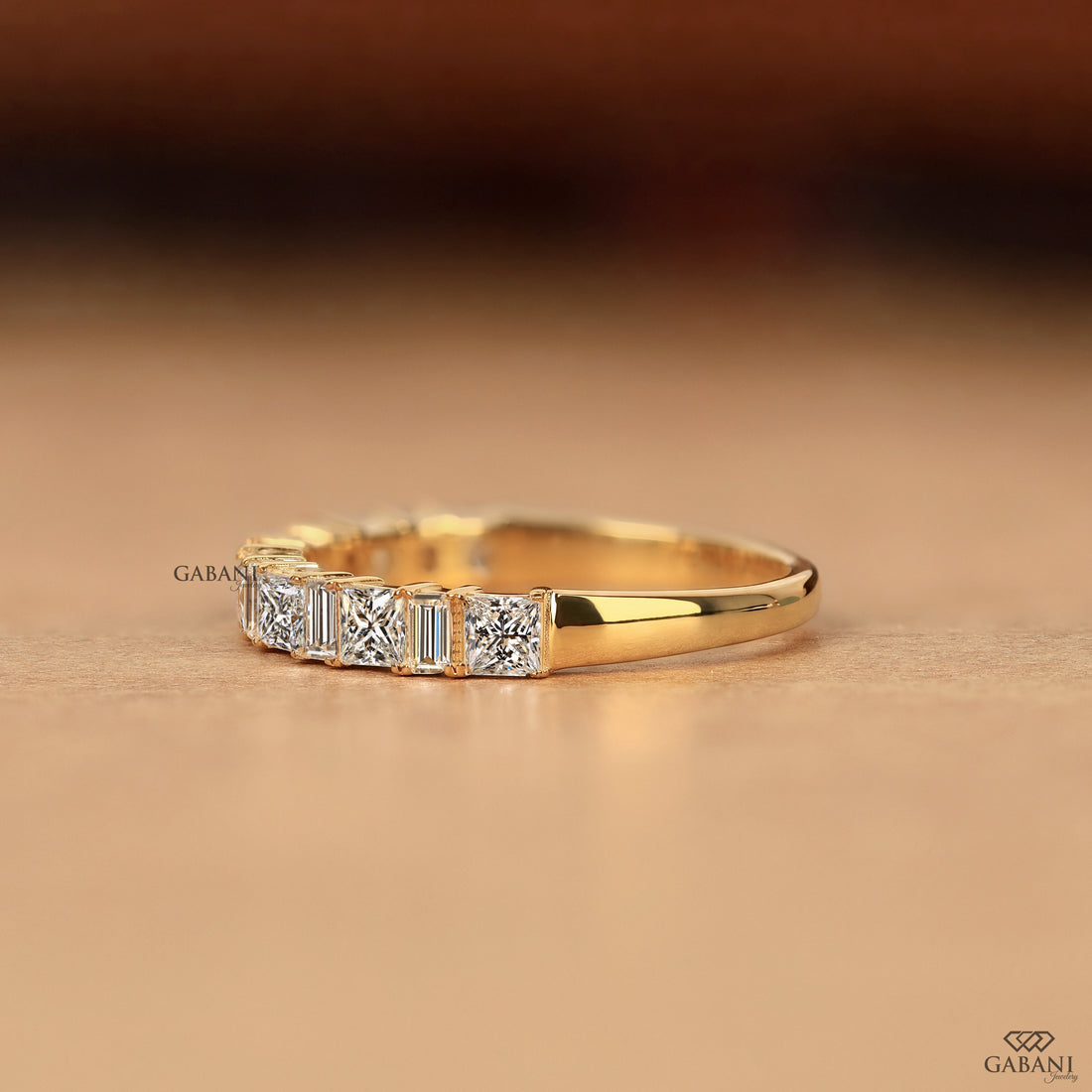 A yellow gold ring featuring a row of baguette-cut and round-cut clear gemstones, displayed on a warm beige surface with a blurred dark brown background.
