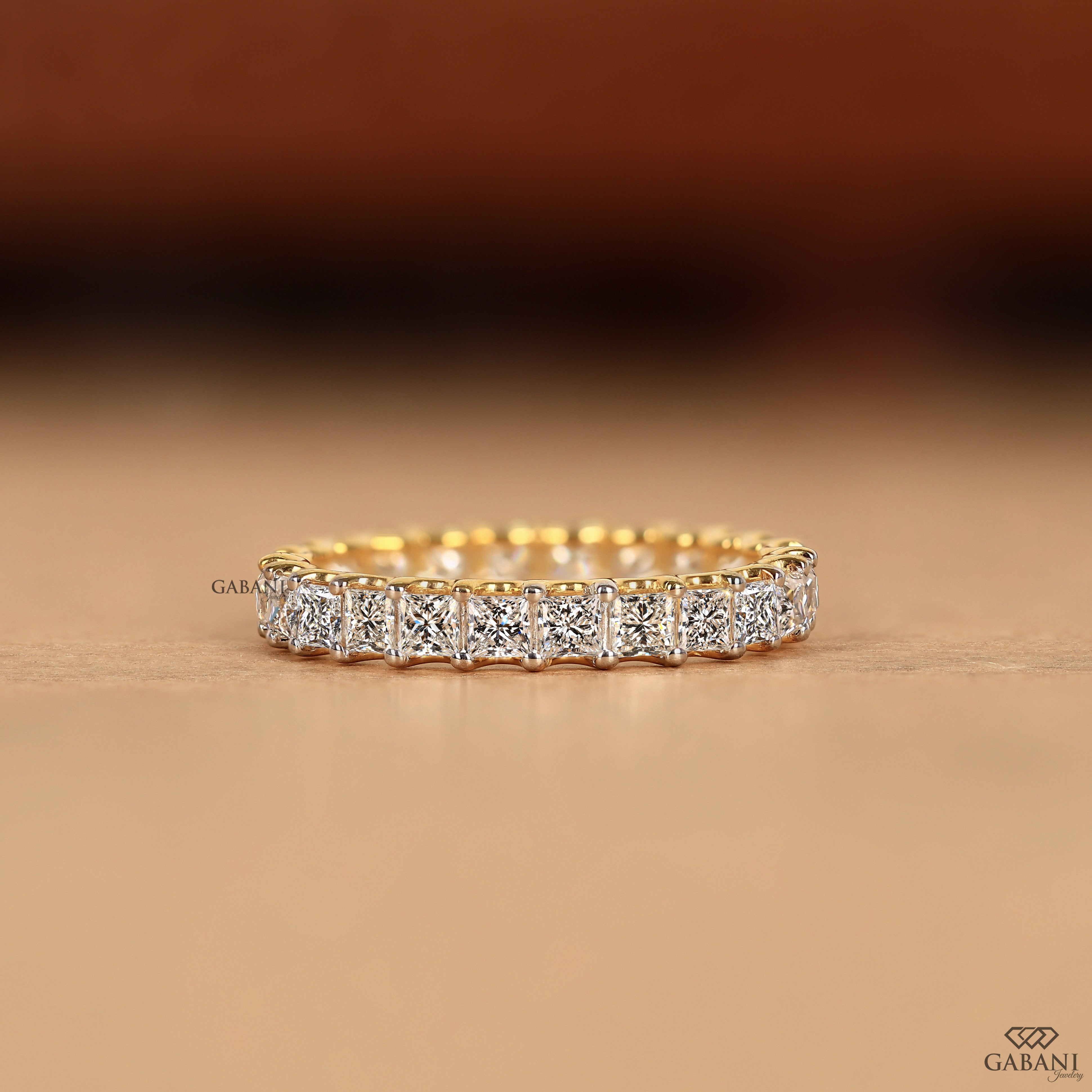 A yellow gold eternity ring featuring a continuous row of square-cut clear diamonds in a bezel setting, displayed on a warm beige surface with a blurred brown background.