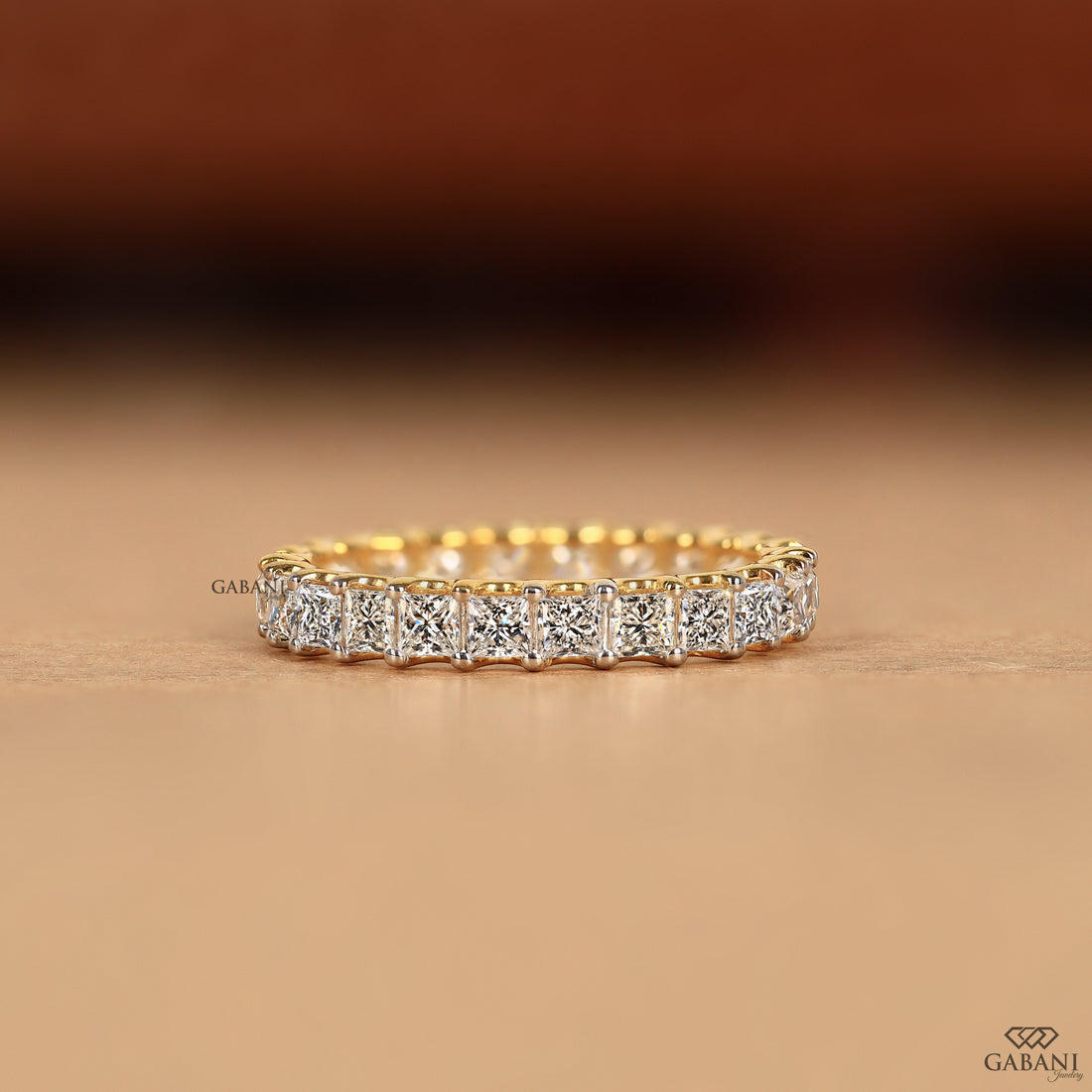 A yellow gold eternity ring featuring a continuous row of square-cut clear diamonds in a bezel setting, displayed on a warm beige surface with a blurred brown background.