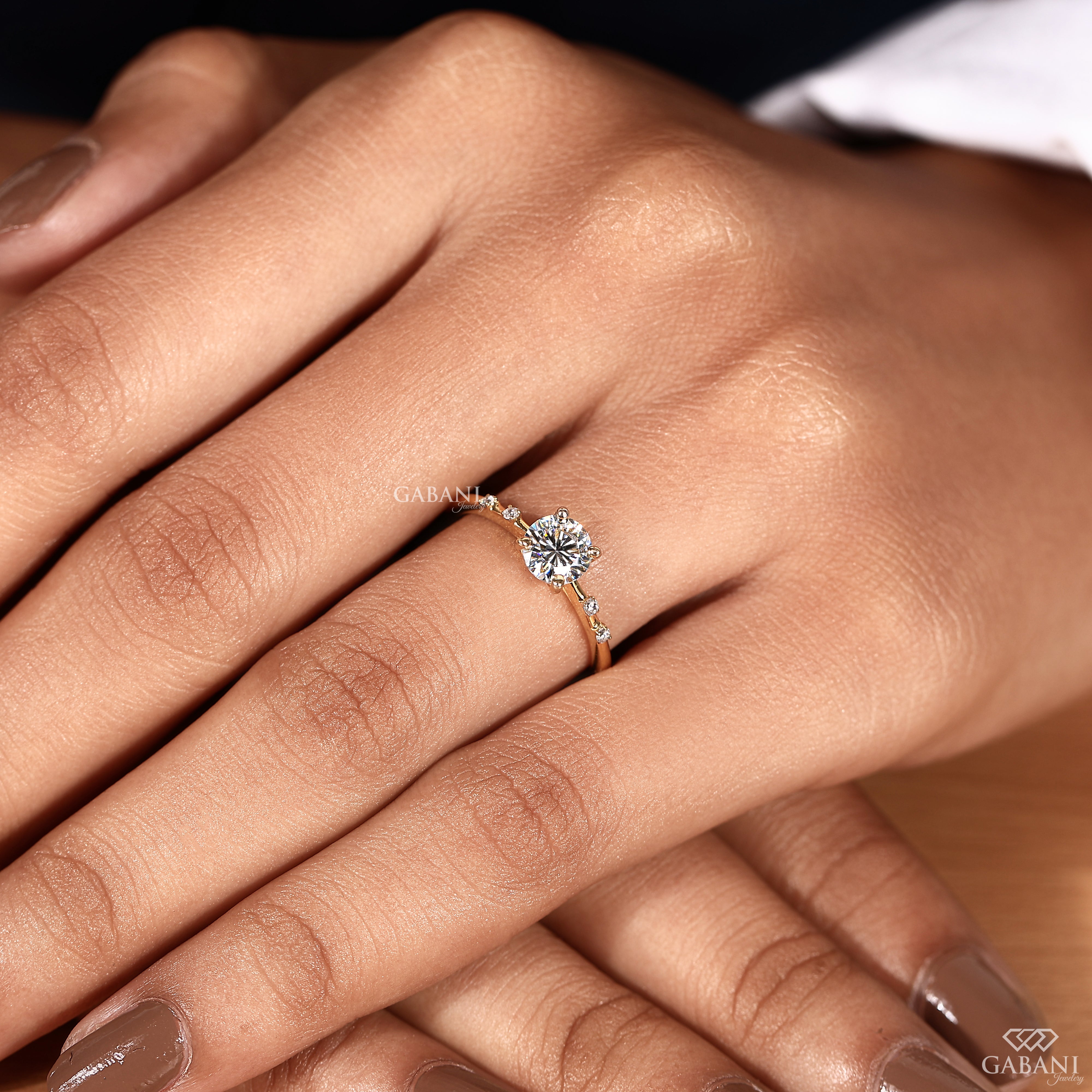 A close-up of a hand wearing a delicate gold ring with a clear round-cut center stone and small side stones, shown against skin tones with a soft blurred background.