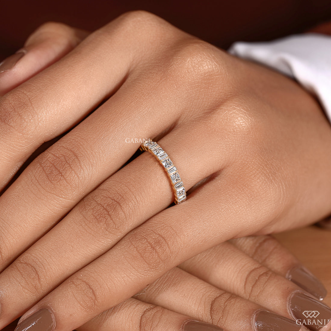 A close-up of a hand wearing a gold band ring set with small clear rectangular stones. The ring is shown on the middle finger against a warm brown background, with a white sleeve visible at the top right.
