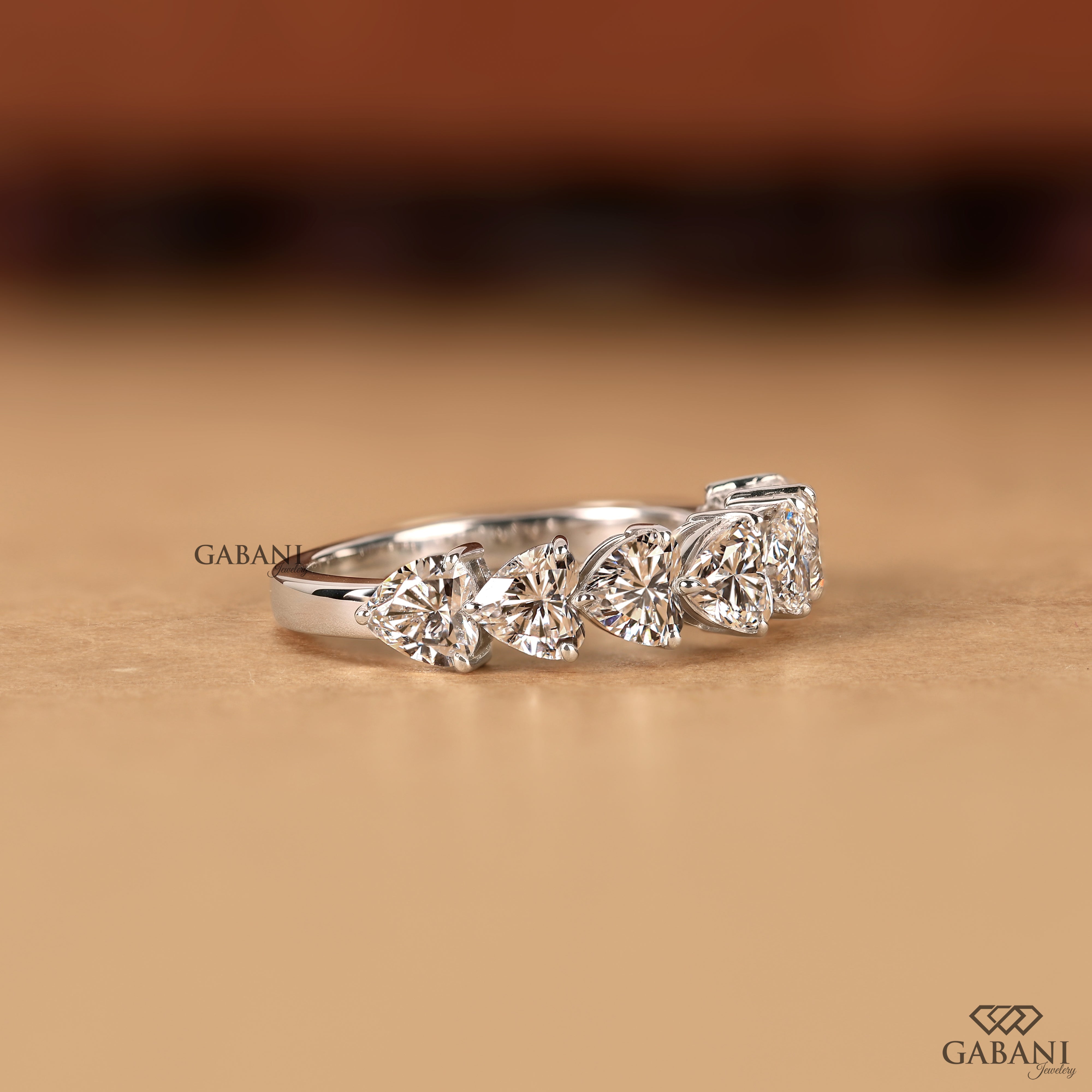 A silver-toned diamond ring with three marquise-cut diamonds in a row, photographed on a warm beige surface with a blurred brown background. The brand name GABANI is visible as a watermark and logo.