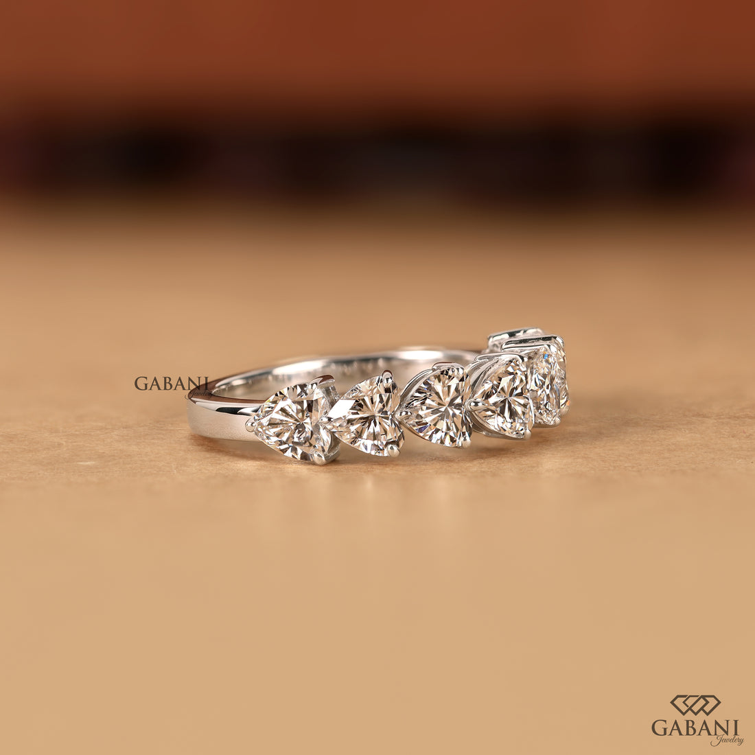 A silver-toned diamond ring with three marquise-cut diamonds in a row, photographed on a warm beige surface with a blurred brown background. The brand name GABANI is visible as a watermark and logo.