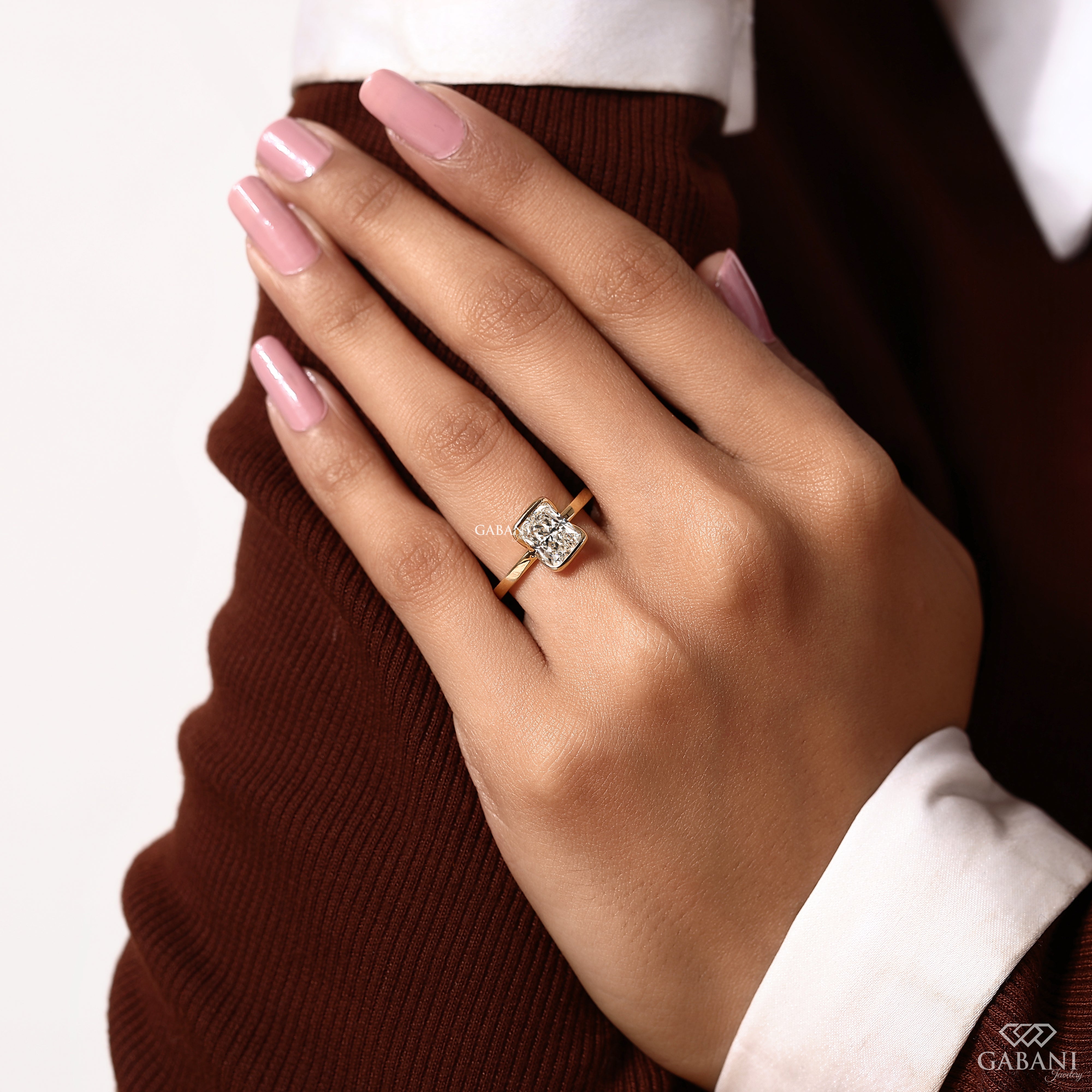 A close-up of a hand wearing a square-cut diamond ring with a rose gold band, styled with a brown knit sweater and white collared shirt. The brand name GABANI is visible in the bottom right corner.