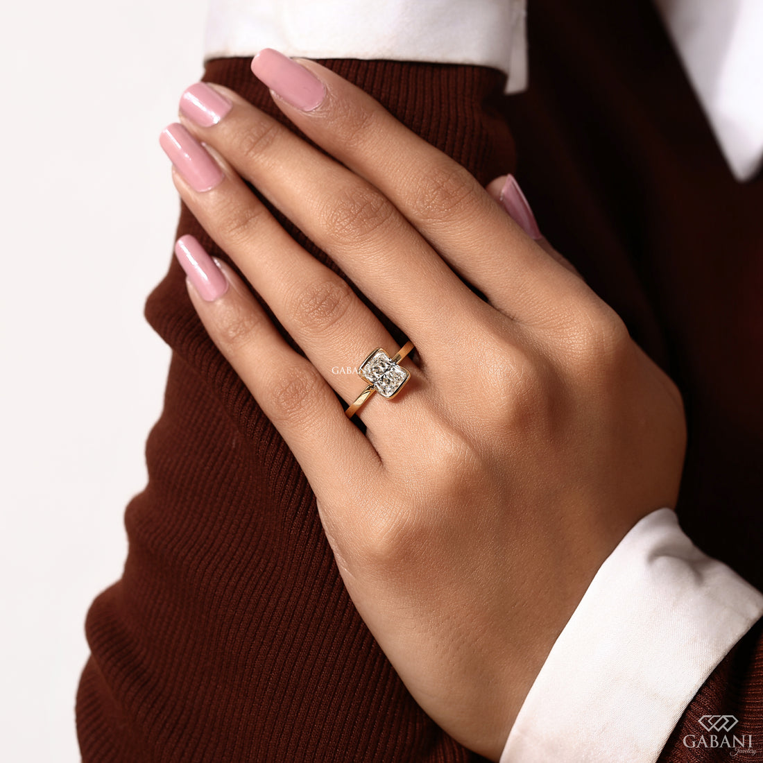 A close-up of a hand wearing a square-cut diamond ring with a rose gold band, styled with a brown knit sweater and white collared shirt. The brand name GABANI is visible in the bottom right corner.