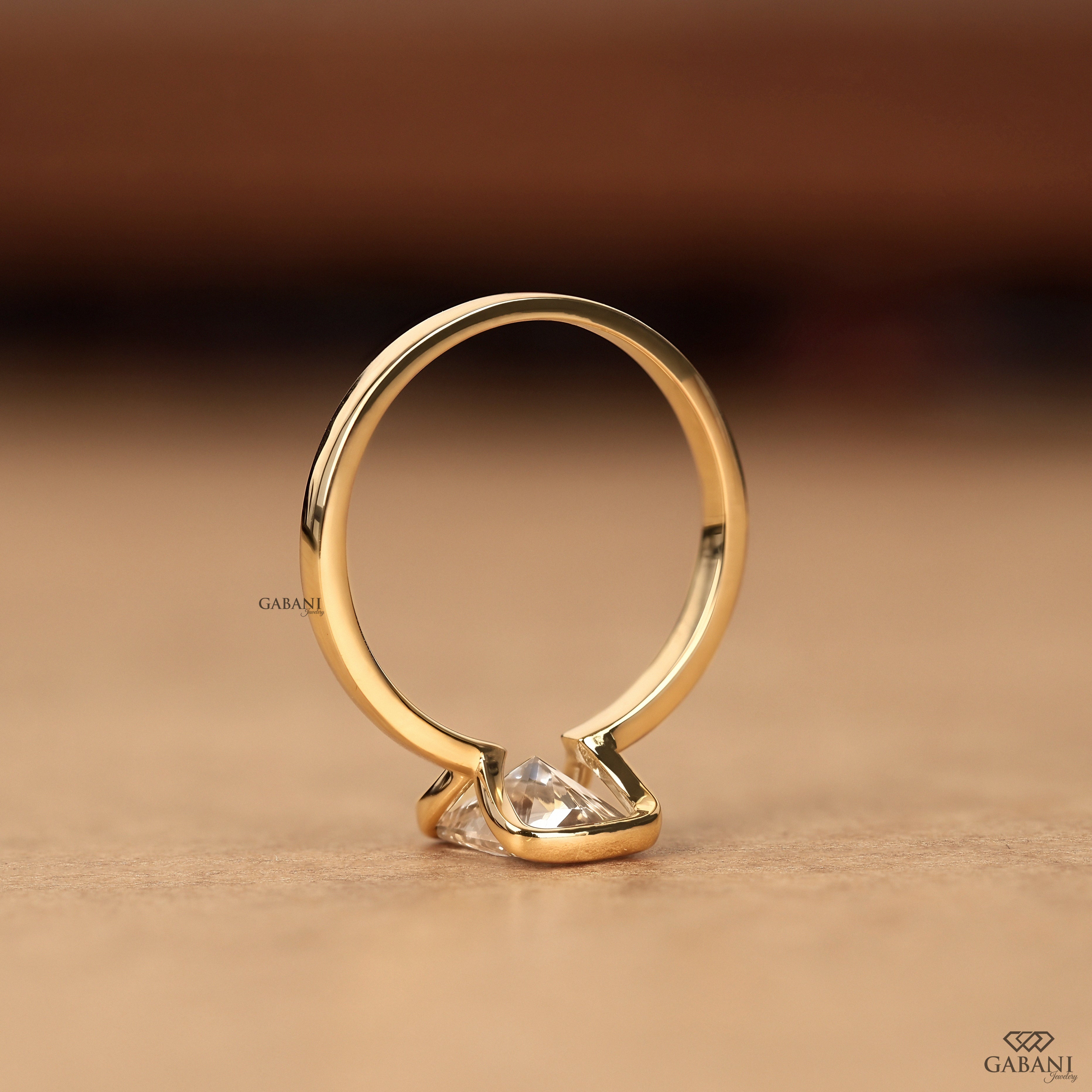 A gold ring with a clear heart-shaped gemstone, photographed on a textured beige surface with a warm, blurred brown background. The brand name 'GABANI' is visible as a watermark and logo.