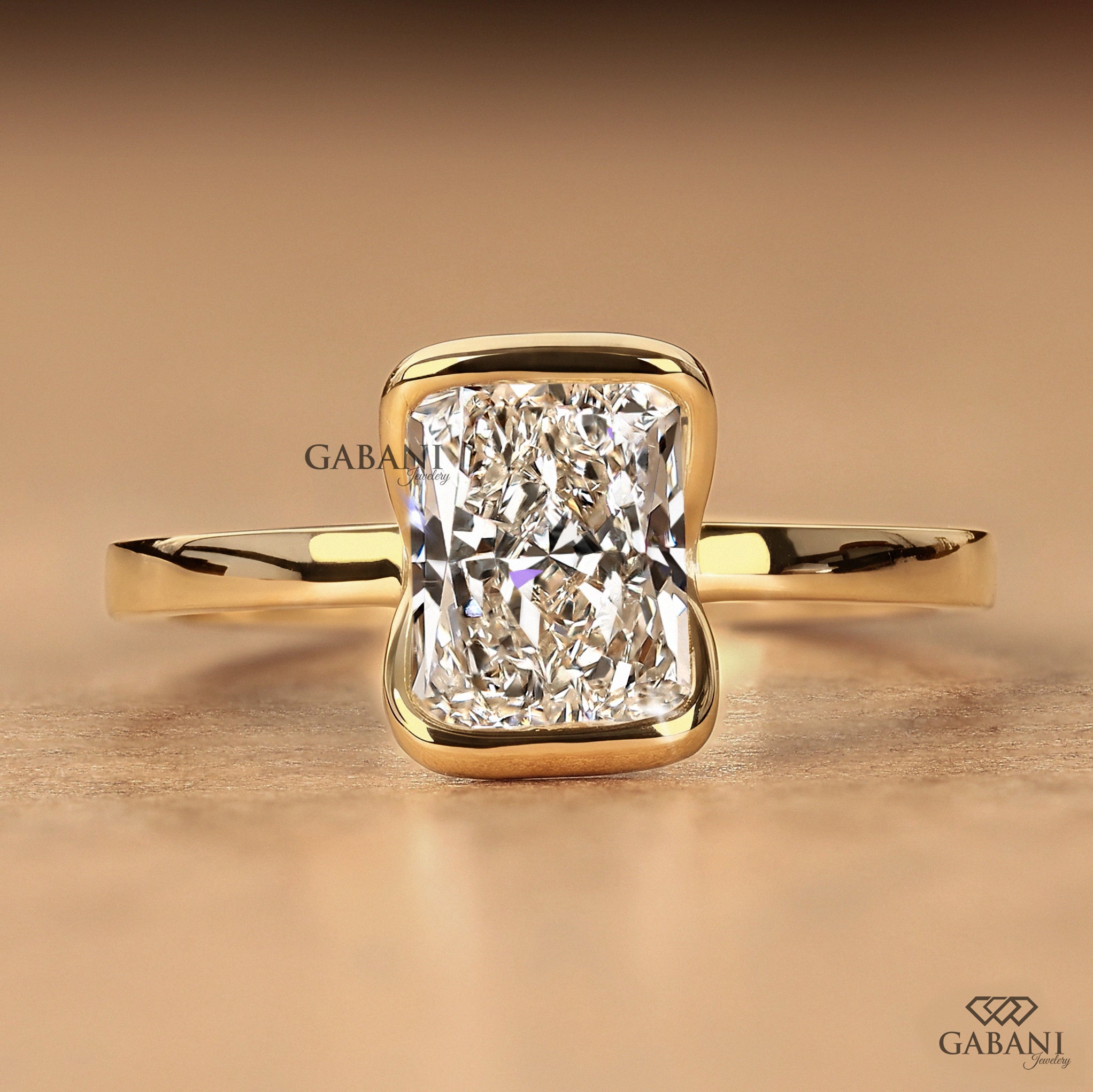 A yellow gold ring featuring a large cushion-cut diamond in a bezel setting, displayed on a warm beige background. The brand name 'GABANI' is visible as a watermark and logo in the lower right corner.