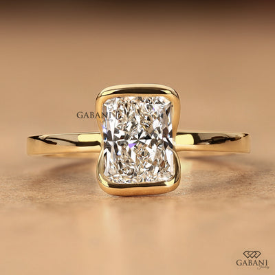 A yellow gold ring featuring a large cushion-cut diamond in a bezel setting, displayed on a warm beige background. The brand name 'GABANI' is visible as a watermark and logo in the lower right corner.