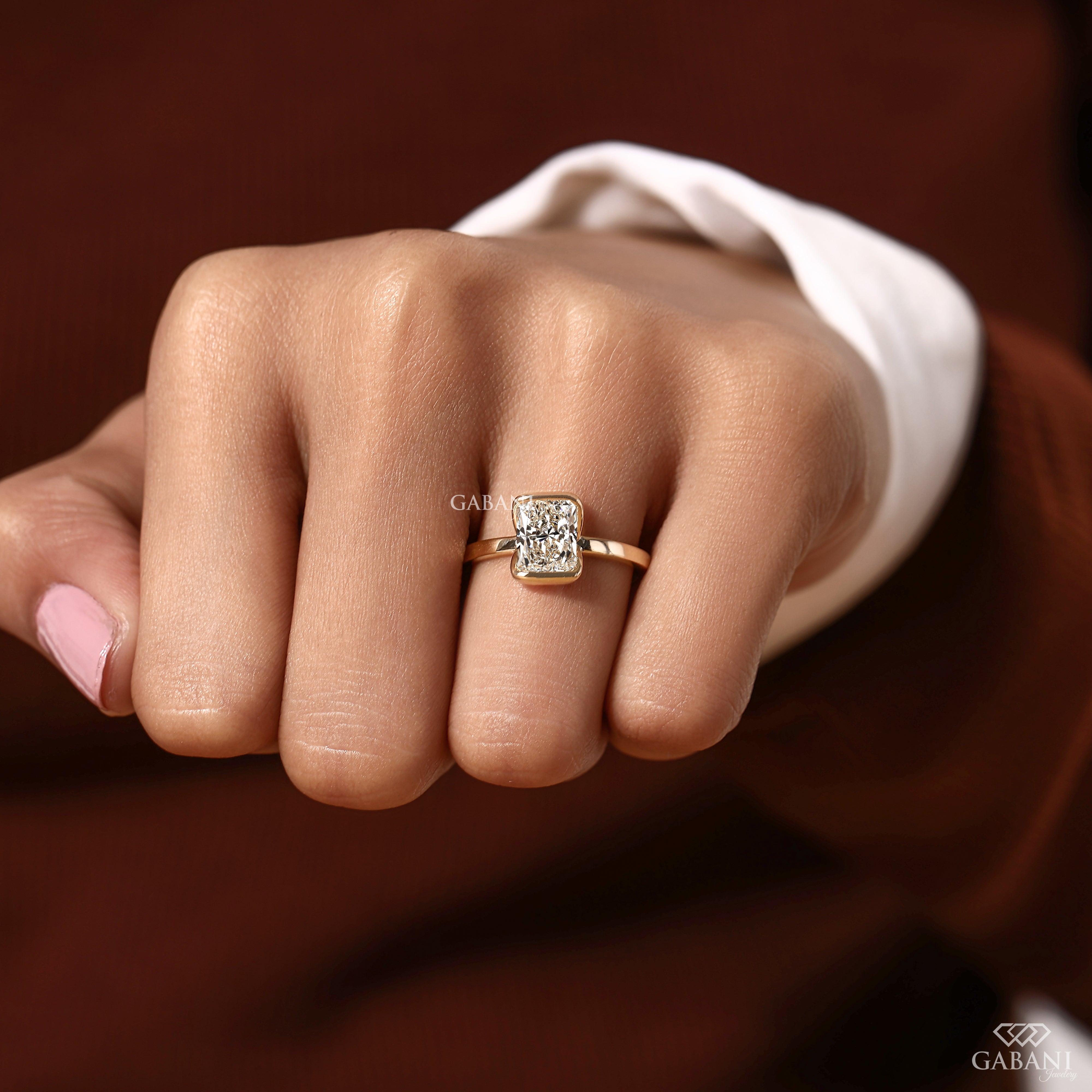 A close-up of a hand wearing a gold ring with a rectangular clear gemstone, set against a dark brown background with a white shirt cuff visible.