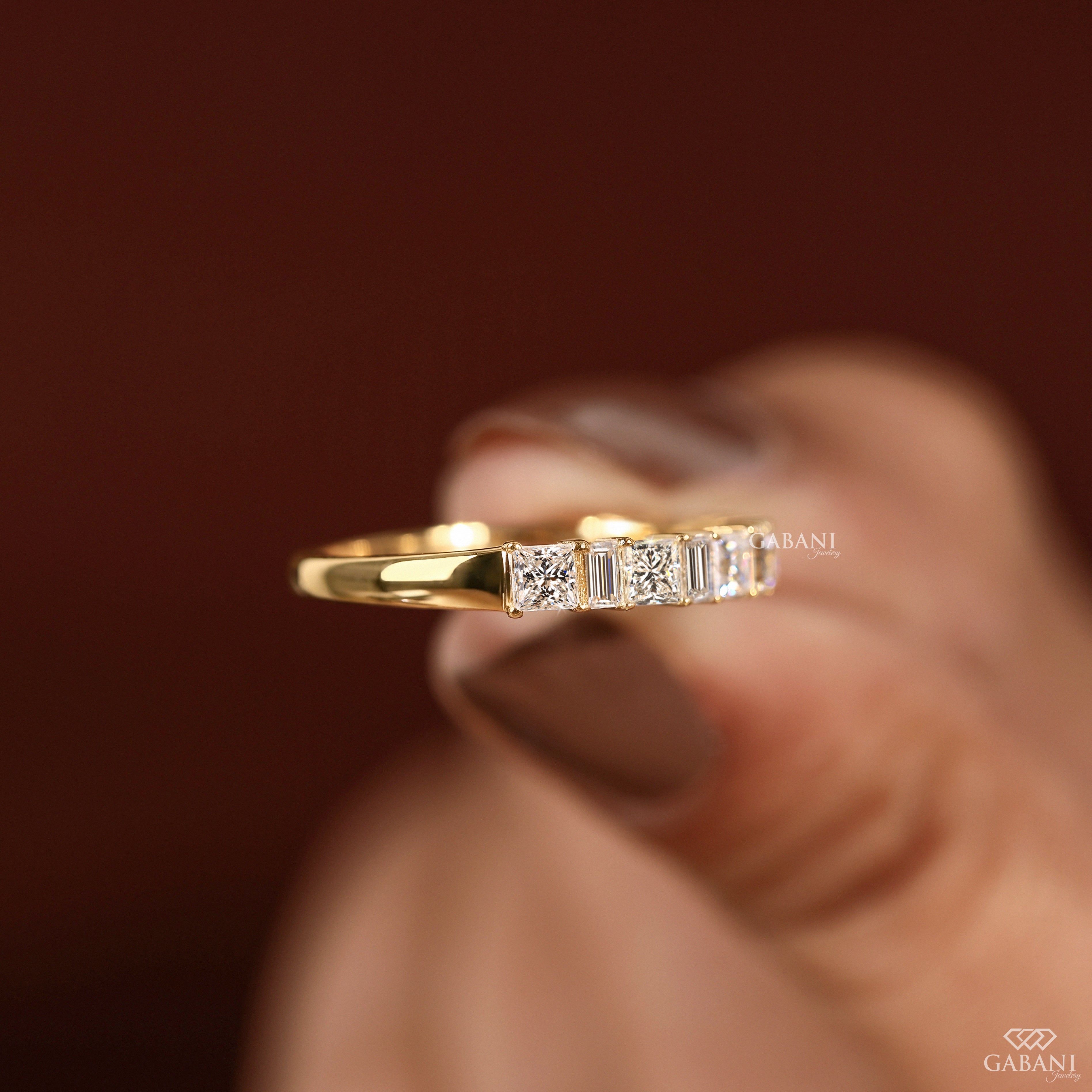 A gold ring with a row of baguette and round-cut clear stones, held between fingers against a dark brown background. The brand name GABANI is visible as a watermark.