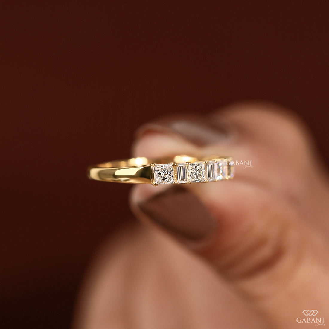 A gold ring with a row of baguette and round-cut clear stones, held between fingers against a dark brown background. The brand name GABANI is visible as a watermark.