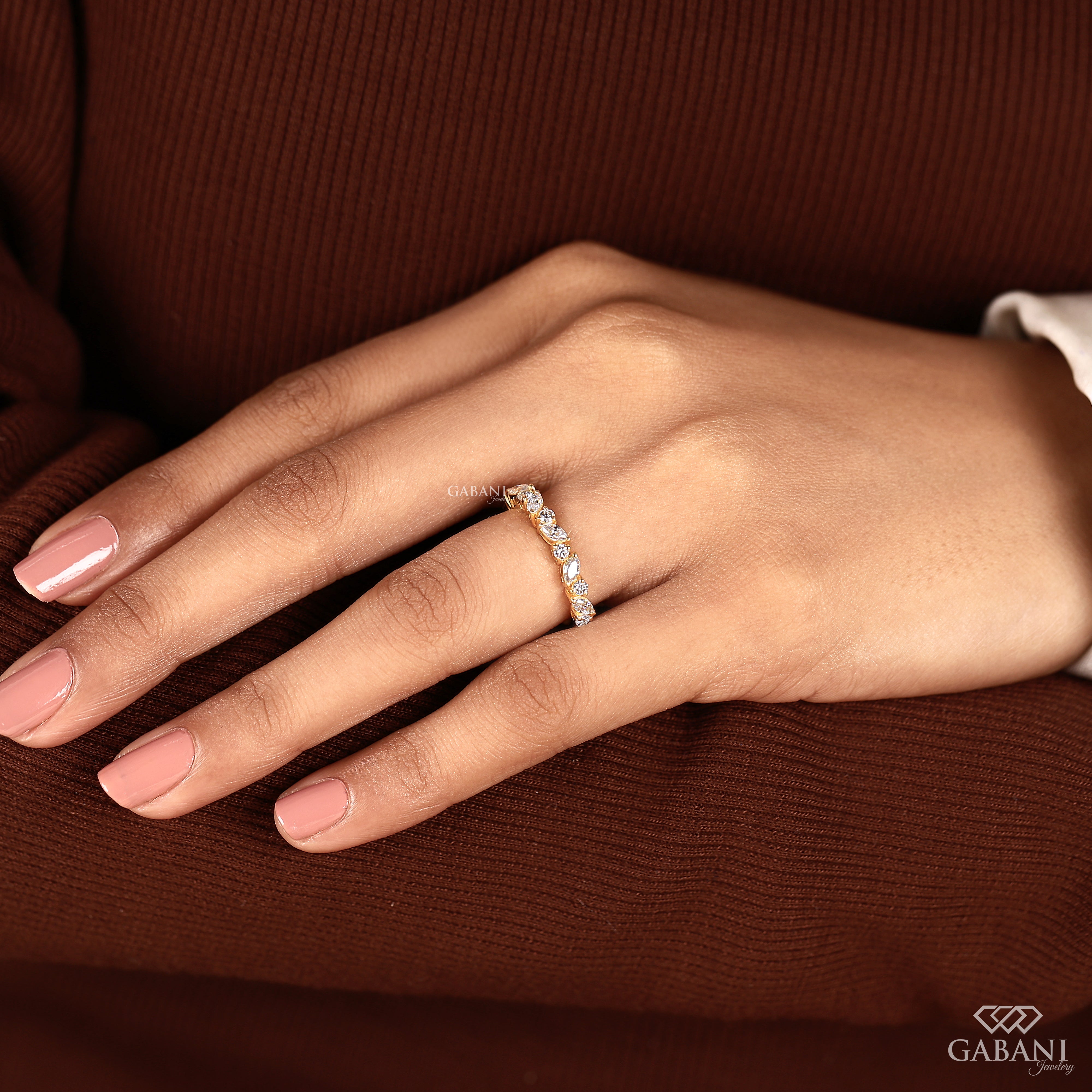 A close-up of a hand wearing a delicate diamond band ring on a warm brown ribbed sweater. The brand name GABANI is visible in the bottom right corner.