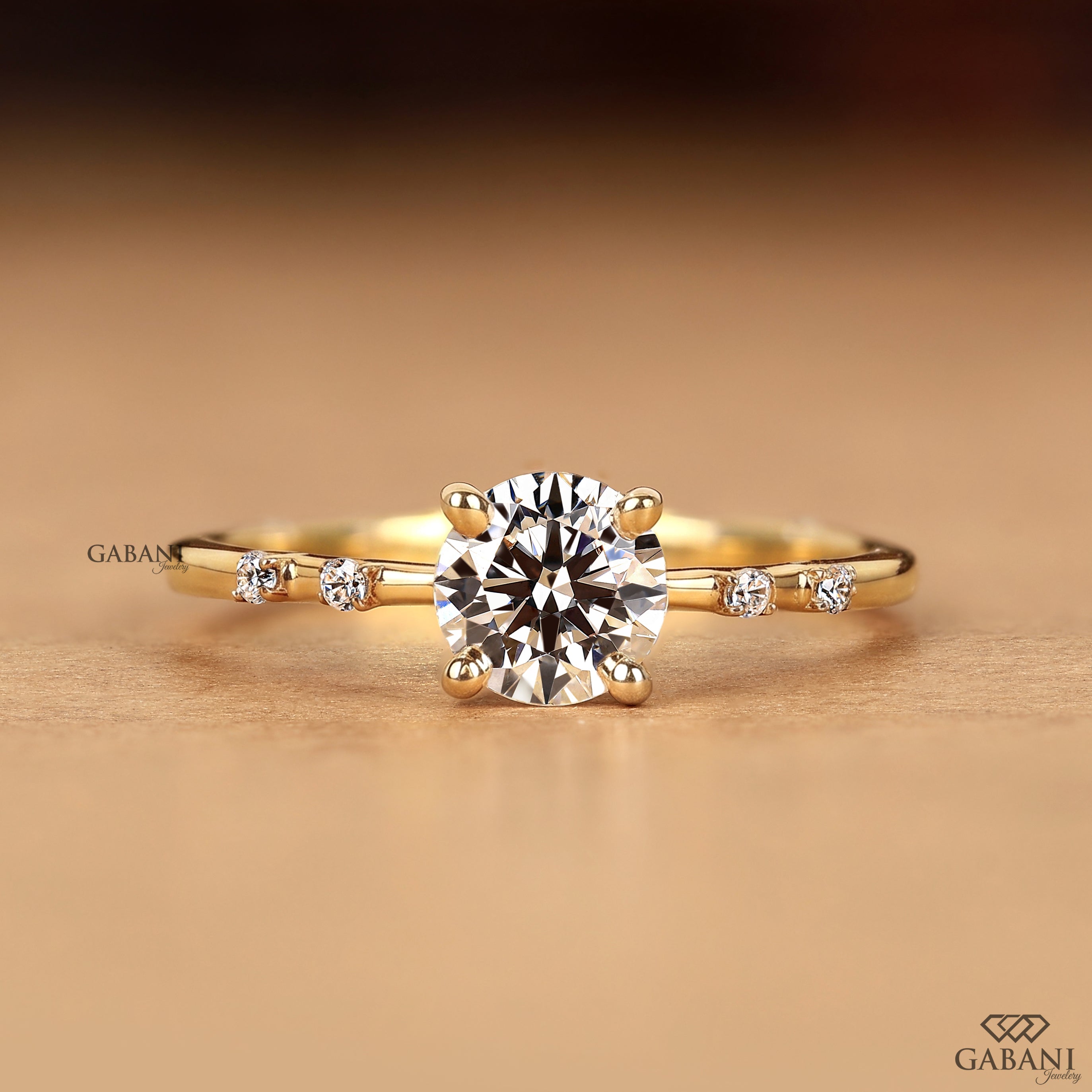 A yellow gold engagement ring featuring a round brilliant-cut center stone and three small side stones on a warm, softly blurred background. The brand name 'GABANI' is visible as a watermark on the left and in the bottom right corner.