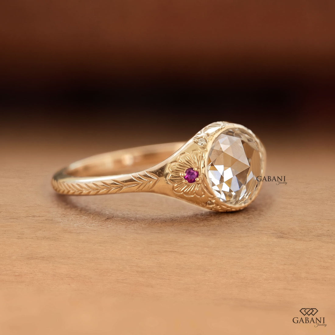 Round rose cut lab grown diamond engagement ring with ruby side accents, floral engraving, and bezel setting in 14K yellow gold.