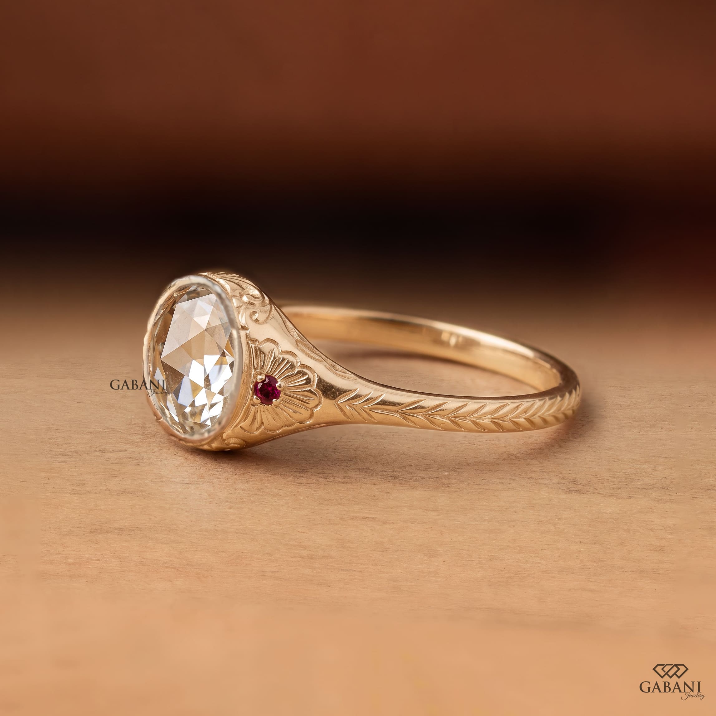 Round rose cut lab grown diamond engagement ring with ruby side accents, floral engraving, and bezel setting in 14K yellow gold.