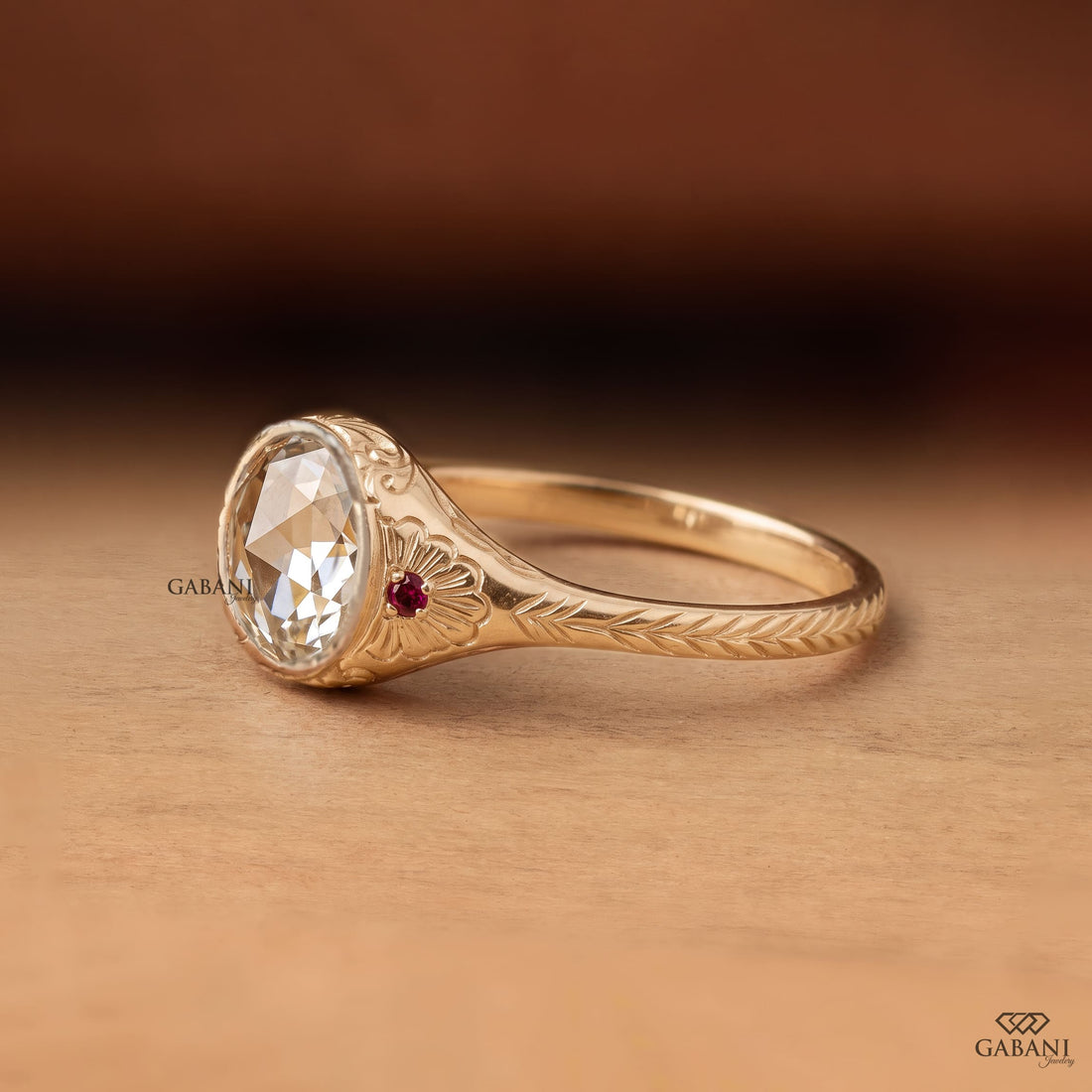 Round rose cut lab grown diamond engagement ring with ruby side accents, floral engraving, and bezel setting in 14K yellow gold.