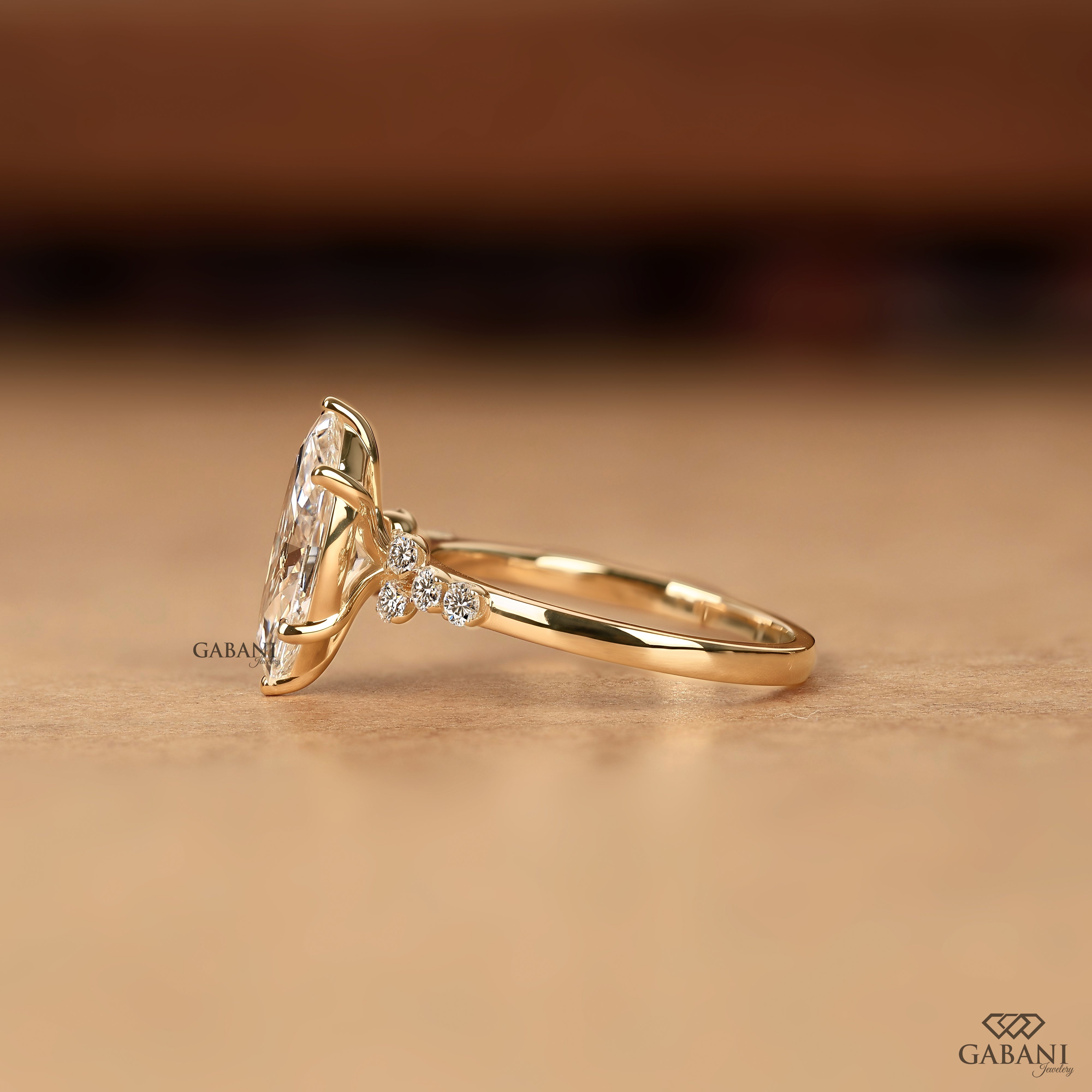 A gold ring with a large marquise clear gemstone and small accent stones on the band, photographed on a warm beige surface with a blurred brown background. The brand name GABANI appears as a watermark and in the bottom right corner.